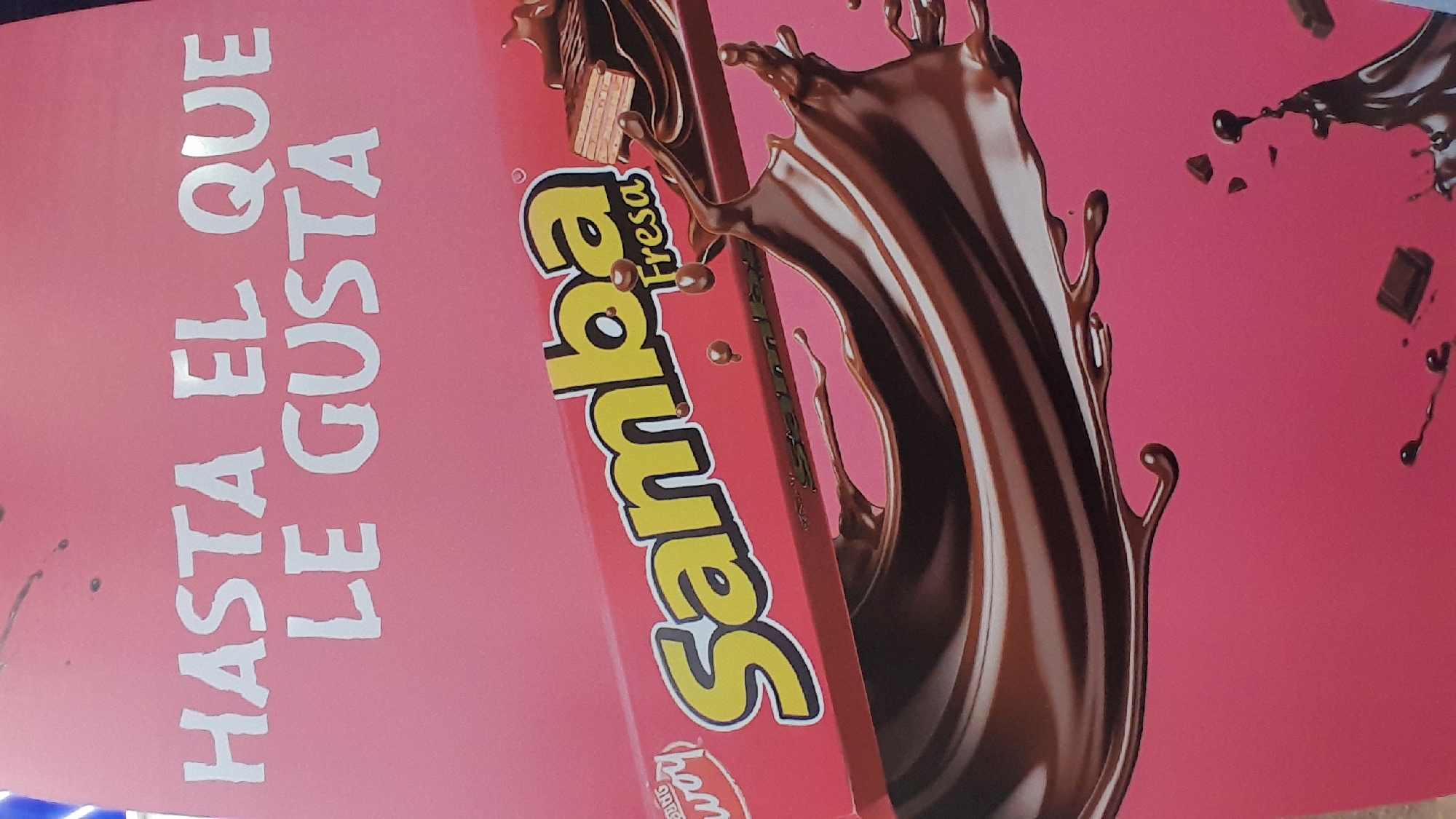 The image is an advertisement for "Samba Fresa" chocolates, displayed on a pink background. The text "HASTA EL QUE LE GUSTA" is prominently displayed in white capital letters. A chocolate bar, labeled "Samba Fresa," is shown partially submerged in splashing chocolate. The advertisement appears to be part of a commercial display, possibly in a store or market in Pampatar, Venezuela. There are no people visible in the image. The scene is static and captures a moment of visual appeal for a confectionery product.