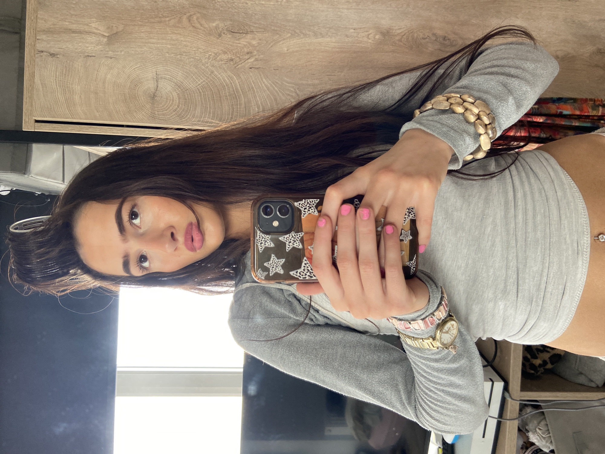 A young woman with long, dark brown hair and pink nail polish is holding a smartphone with a patterned case, presumably taking a selfie. She is wearing a fitted, light gray long-sleeved top, a gold bracelet on her left wrist, and a wider, chunky gold bracelet on her right arm. A gold watch is visible on her left wrist as well. She is positioned indoors with a light-colored wooden surface behind her and what appears to be a window to her left, indicating natural light. The background also shows parts of furniture and other items, suggesting a casual indoor setting. It is not possible to determine the specific location within London, United Kingdom from the provided image.