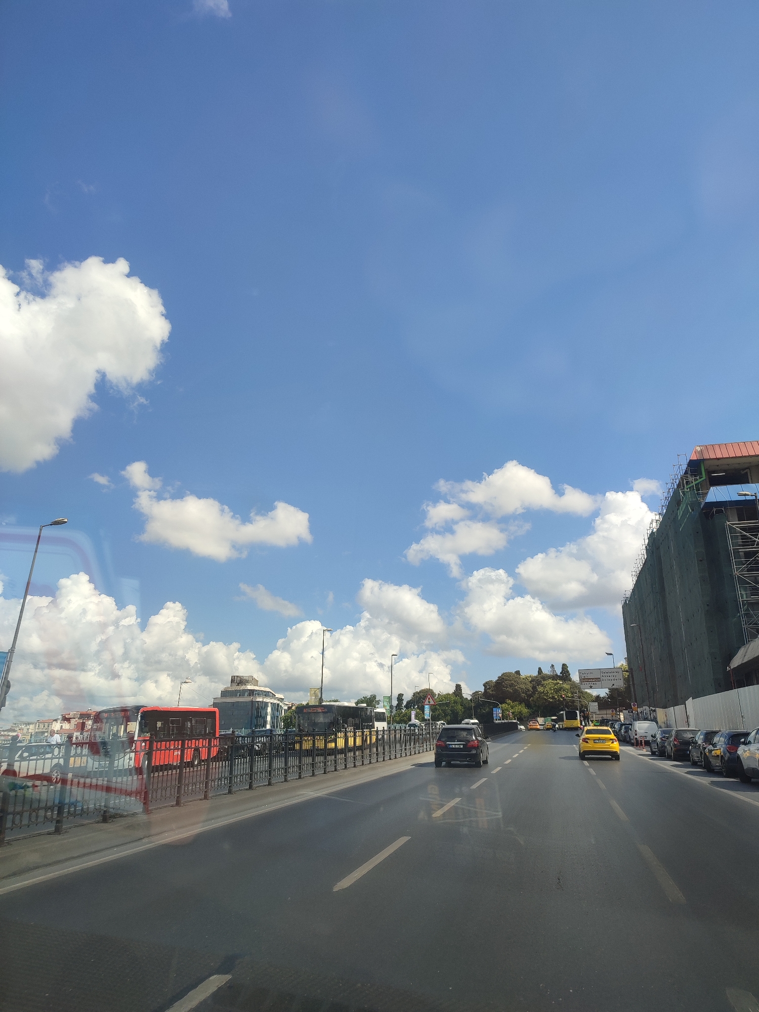 The image shows a street scene in Beyoğlu, Turkey, on a clear day with scattered white clouds. A multi-lane road dominates the foreground, with vehicles including cars, buses, and a taxi. On the left side of the road, there is a sidewalk and a black metal fence separating it from a pedestrian walkway. Beyond the walkway, several buses, including a red one and a white one, are visible. A tall building, partially covered in scaffolding and green netting, stands on the right side of the road. Trees and more buildings can be seen in the background. A street lamp is visible on the left. The overall impression is of a busy urban environment under a bright blue sky.