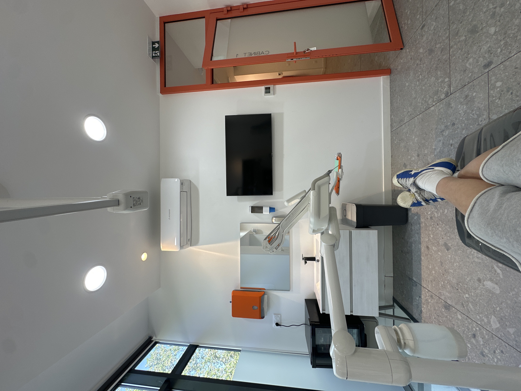 The image shows the interior of what appears to be a dental office. The room has white walls, with several fixtures including a flat-screen TV mounted on the wall, an air conditioning unit, a sink, and a cabinet. The dental equipment is visible, including a dental light and other apparatus. A person is sitting in the dental chair. The scene includes an orange framed doorway and a window with a view of trees outside. It is not possible to determine the precise location within Paris from the image alone.