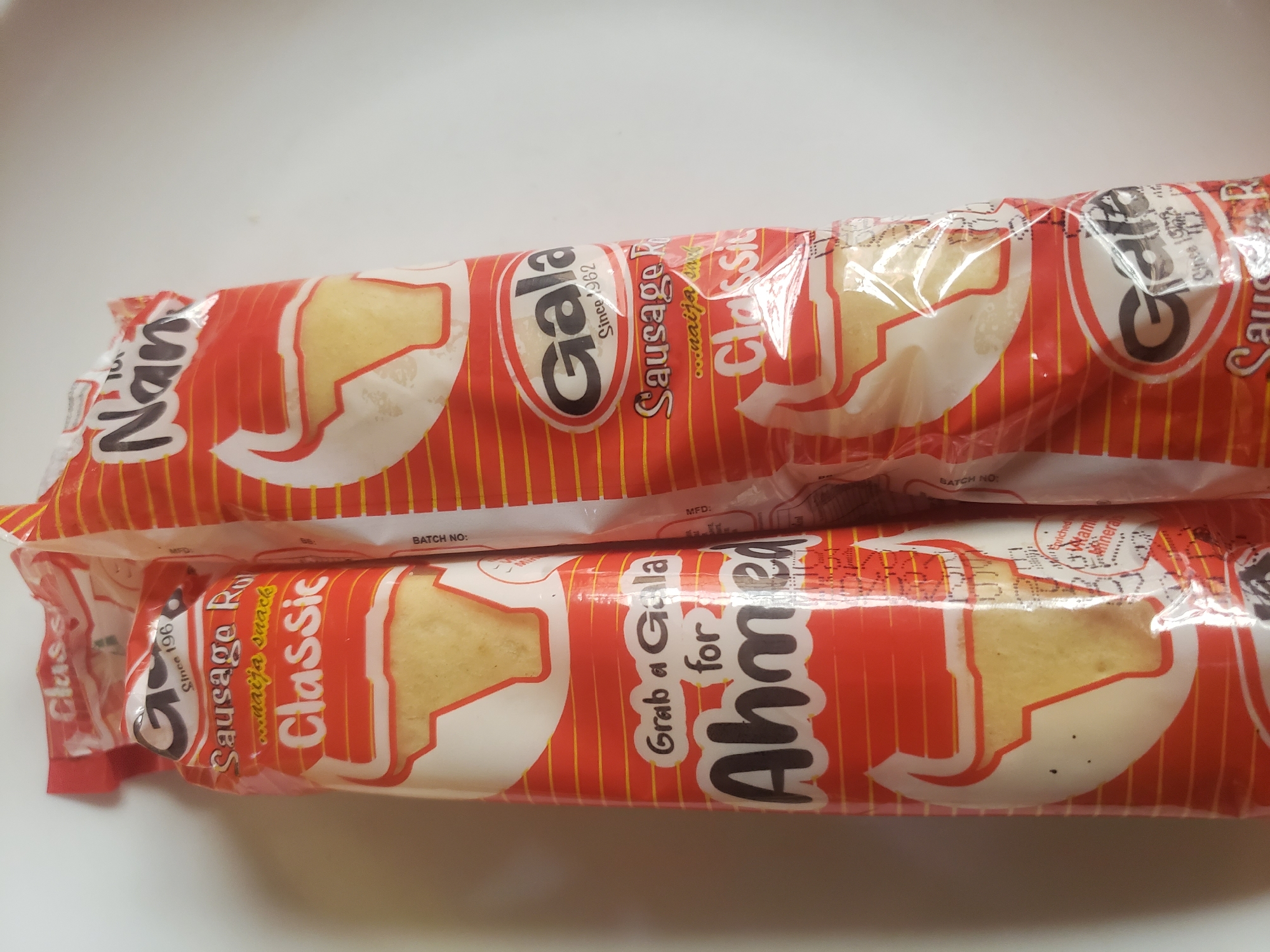 Two packages of Gala sausage rolls are displayed on a white surface. The package on the left is partially obscured by the package on the right. Both packages feature a red and orange striped design with a white oval containing a bull's head and a portion of the sausage roll. Text on the packages includes "Gala", "Since 1962", "Sausage R...", "...naija snack", and "Classic". The package on the left also has the text "Grab a Gala for Ahmed". The packages appear to be in good condition with no visible damage. The environment is a plain white background, suggesting a studio or indoor setting. There are no people or other objects present in the image. The lighting is even and appears to be from overhead.
