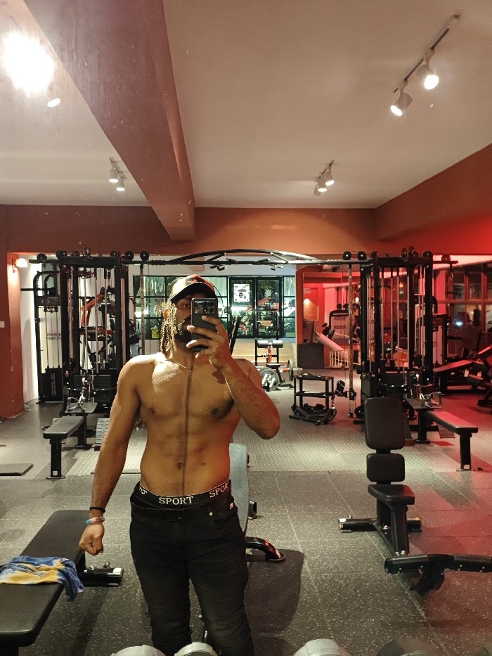 A shirtless man in a gym takes a mirror selfie with his phone. He wears black pants with a "SPORT" waistband and a baseball cap. Gym equipment and weights surround him, including weight machines and benches. The gym's interior features grey floors, red accent lighting, and a mirrored wall. Overhead lights illuminate the scene.