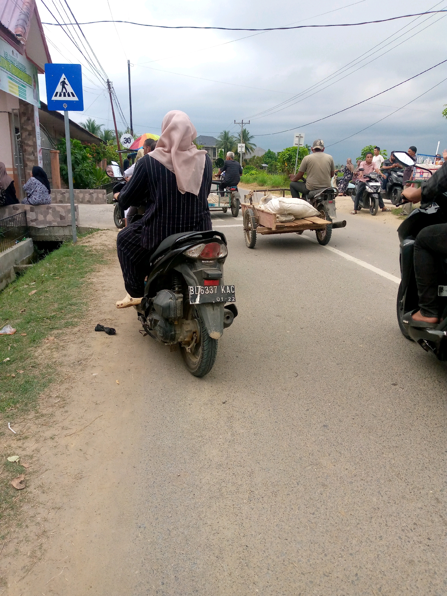 A busy street scene in Mesjid, Indonesia shows a variety of motorbikes and pedestrians. The focus is on a woman wearing a pale pink hijab and a black and white striped abaya, riding a black Suzuki motorbike. Her right foot is extended, and a pair of black sandals are placed on the ground next to the motorbike. The motorbike's license plate is visible and reads "B U6337 KAC 01 22". Behind her, a man on another motorbike pulls a small, rustic trailer carrying a white sack. In the background, several other motorbikes and pedestrians are visible, with power lines crisscrossing the overcast sky. A blue pedestrian crossing sign is prominently displayed on the left side of the road. The ground is a mix of asphalt and dirt.