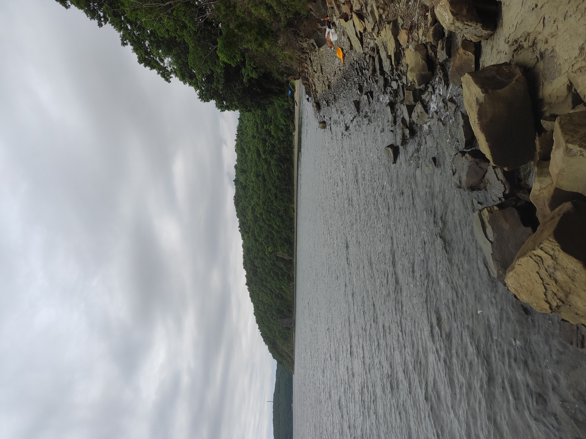 The image depicts a rocky shoreline along a body of water, likely the sea, under an overcast sky. A dense, green forest covers the rolling hills that recede into the distance. In the foreground, large, rough-hewn rocks are scattered along the water's edge, with smaller pebbles and debris filling the gaps. The water itself is choppy and dark grey.
Further along the shore, a small sandy beach is visible, leading to more trees. On the right side of the image, several people are present near the shore, with what appears to be camping gear, including a bright orange tarp and a white cooler. There are two individuals walking on the distant beach.
The scene is located on Russky Island, near the city of Vladivostok, Russia. The island is known for its natural beauty, with numerous bays, beaches, and forested hills, making it a popular destination for outdoor recreation and tourism. The presence of the rocks, water, and forest are characteristic of the island's landscape.
