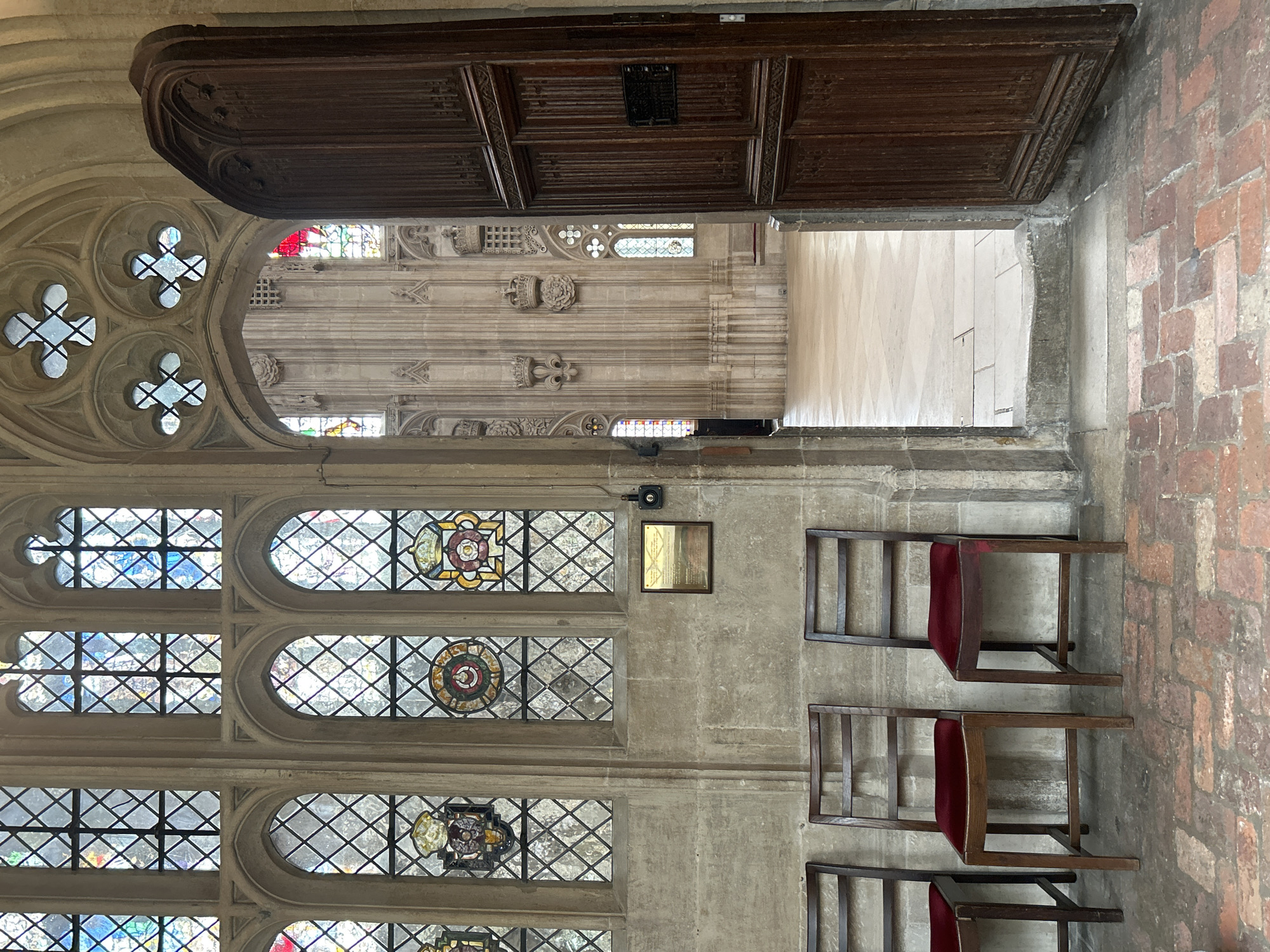 The image depicts an interior view of a historic building, likely a church or cathedral, in Cambridge, United Kingdom. In the foreground, three dark wooden chairs with red velvet seats are lined up against a stone wall. Above the chairs, a large stained-glass window with diamond-shaped panes and decorative crests is visible. To the right, a tall, ornately carved wooden door is open, revealing a glimpse into another part of the building. This inner space features tall, ribbed stone walls adorned with crowns and crests, and more stained-glass windows. The floor in the visible interior is tiled in a diamond pattern. A small plaque is mounted on the stone wall to the right of the chairs. The overall impression is one of architectural grandeur and historical significance.