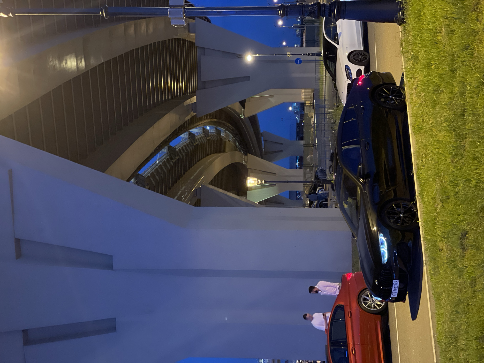 An eye-level, outdoor, medium shot captures a street scene at dusk under a large, multi-tiered overpass. The overpass's concrete supports dominate the left side of the frame, casting shadows across the road. To the right, a black BMW sedan with illuminated blue headlights and license plate "V777EE 178" is parked on the road, partially obscuring a white Mercedes-Benz sedan behind it. Two men in casual attire are standing near a red sedan on the left side of the road, one holding a phone. The sky is a deep twilight blue, with some distant lights visible. The foreground features a strip of green grass. A streetlamp with a yellow glow stands to the right, and other streetlights are visible further back, adding to the ambient lighting of the scene.