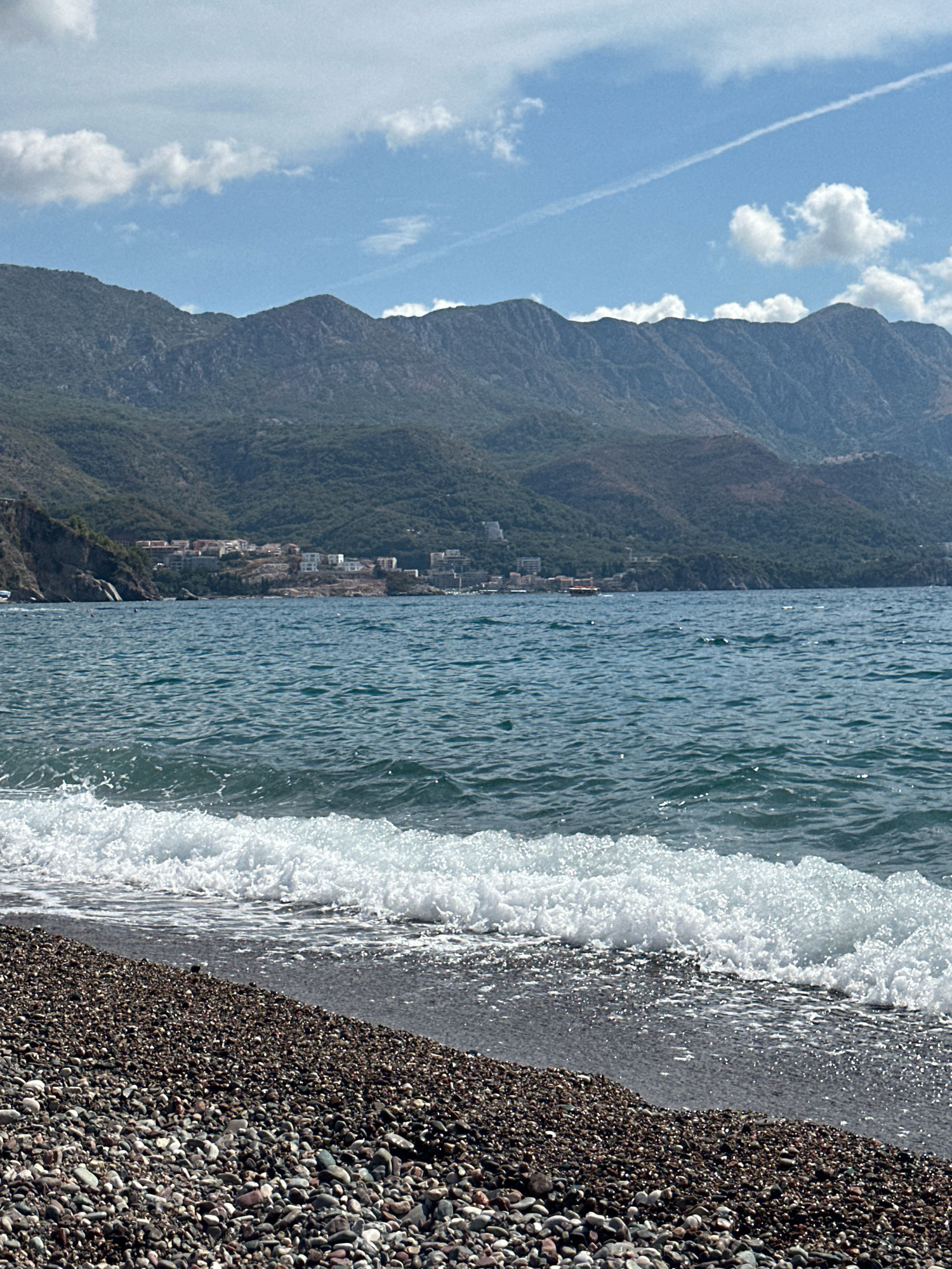 The image captures a serene coastal scene in Boreti, Montenegro. A pebble beach occupies the foreground, with a gentle wave breaking at its edge, creating a foamy white crest that recedes into the dark, wet pebbles. The sea, a deep blue, stretches towards the horizon, dotted with small ripples and glints of sunlight. In the mid-ground, a cluster of buildings, likely a resort or town, is nestled against the base of lush, green mountains that dominate the background. The mountains rise steeply, their slopes covered in dense vegetation, fading into a hazy blue at their peaks. The sky above is a clear blue, punctuated by scattered white clouds, and a faint contrail from an airplane stretches across the upper right quadrant. The overall impression is one of natural beauty and tranquility, with a hint of human habitation integrated into the landscape.