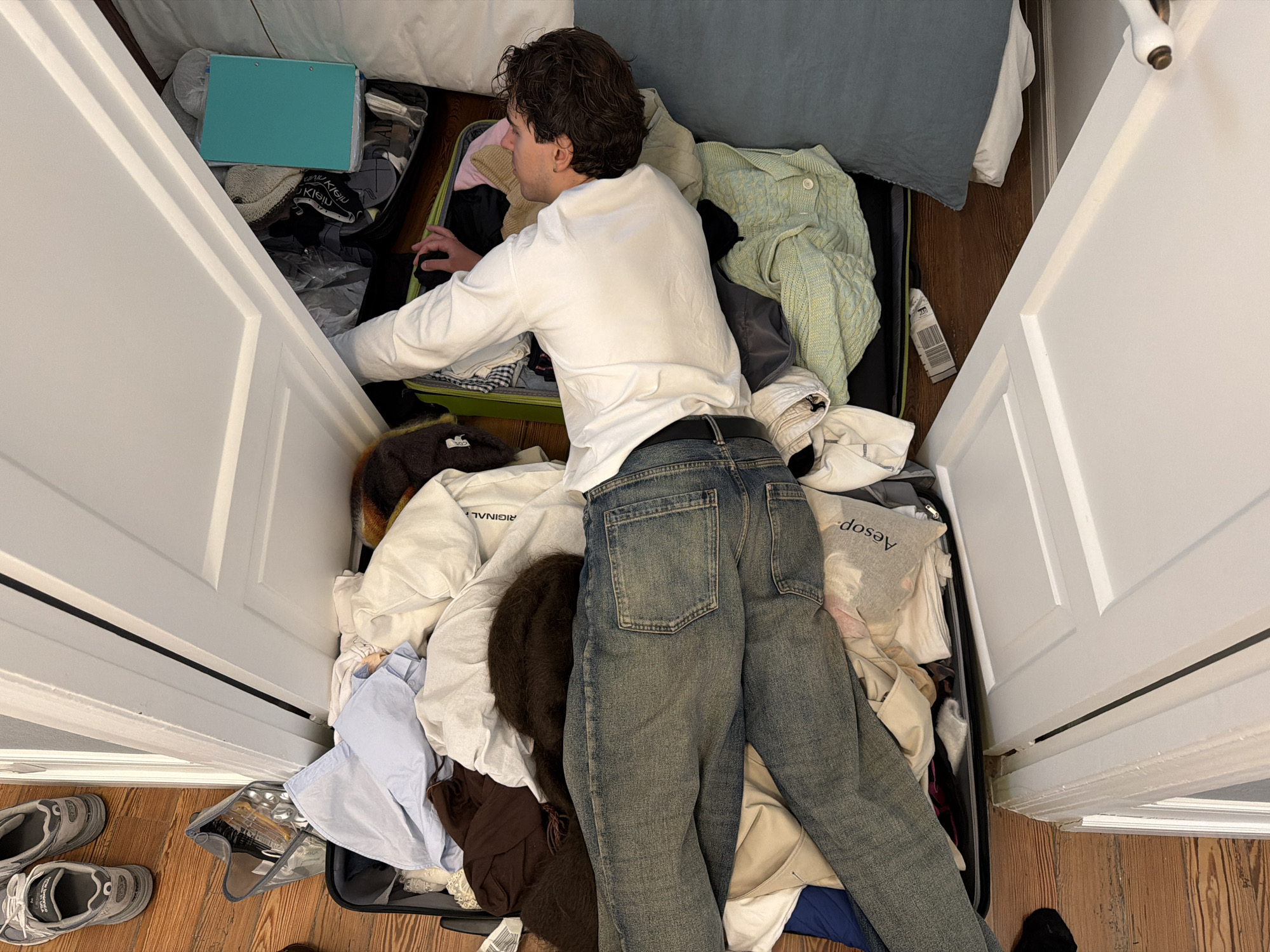 A person wearing a white long-sleeved shirt and blue jeans is lying on top of clothing and suitcases in a closet. The closet doors are open. There are two open suitcases and a turquoise folder on top of the clothes. A pair of grey sneakers is on the floor next to the closet. The room appears to be indoors and well-lit. The location is not discernible as Santiago De Compostela, Spain based on visual cues.