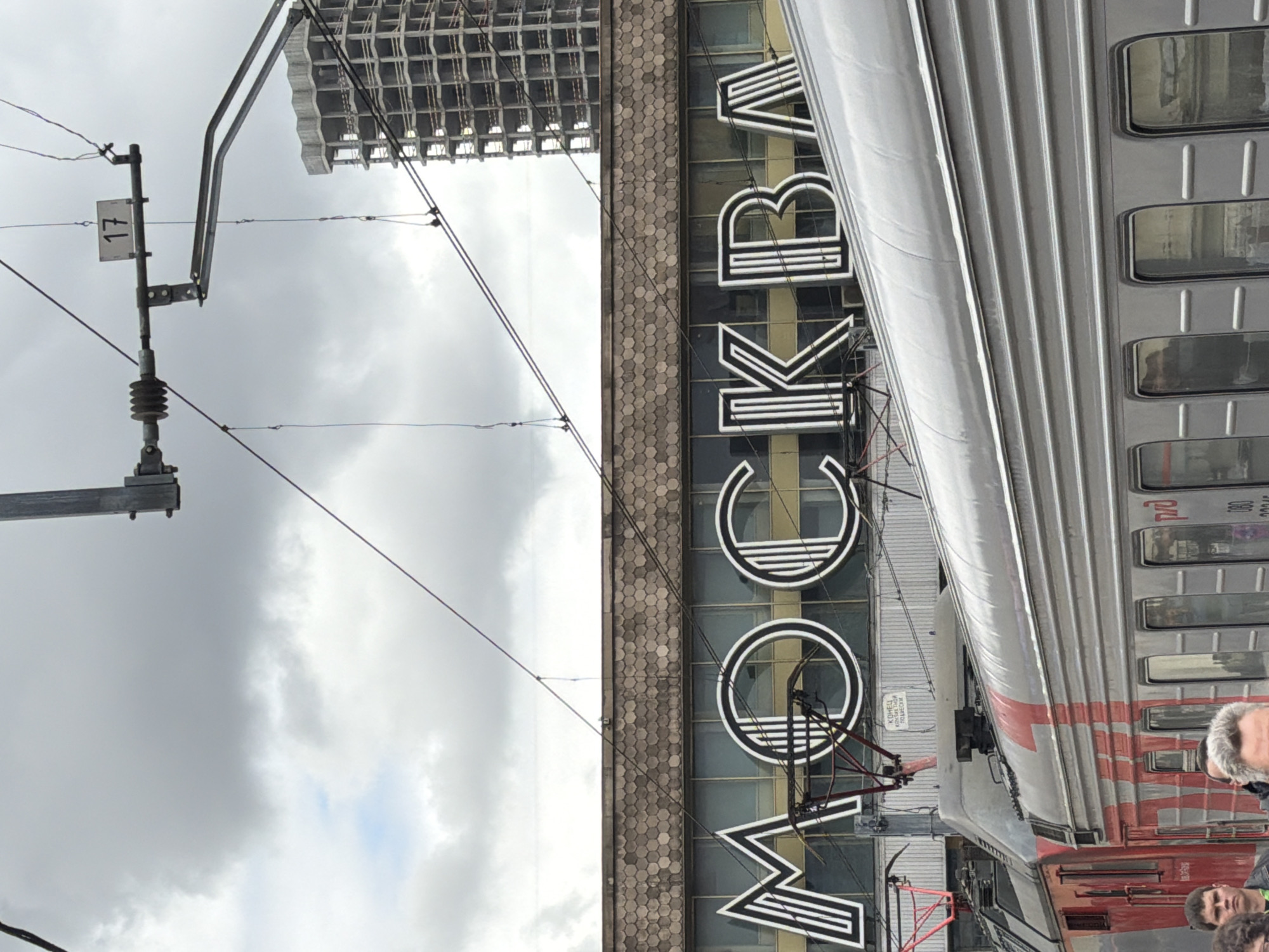 A daytime photograph depicts a train parked alongside a building at what appears to be a railway station in Moscow, Russia. The sky is overcast and cloudy, suggesting a dull day.
In the foreground and left, a railway catenary system with overhead power lines and support structures is prominent. A white sign attached to a pole displays the number "17". Further along the tracks, another sign reads "КОНЕЦ КОНТАКТНОЙ СЕТИ" (End of Contact Network).
On the right side of the image, a silver train carriage is visible, featuring multiple rectangular windows. The logo "РЖД" (RZD, Russian Railways) is printed on its side. Part of a red locomotive or another carriage is seen at the bottom right. Two individuals are partially visible at the bottom right, near the train, one with gray hair and another looking forward.
Behind the train and spanning the center-left is a building facade with large windows. A prominent, large sign spelling "МОСКВА" (MOSKVA) in stylized, outline Cyrillic letters is affixed to this building. Below the sign, the building's facade features a textured pattern of hexagonal tiles. Above this, a large, partially constructed or derelict building with a concrete grid structure rises against the sky.