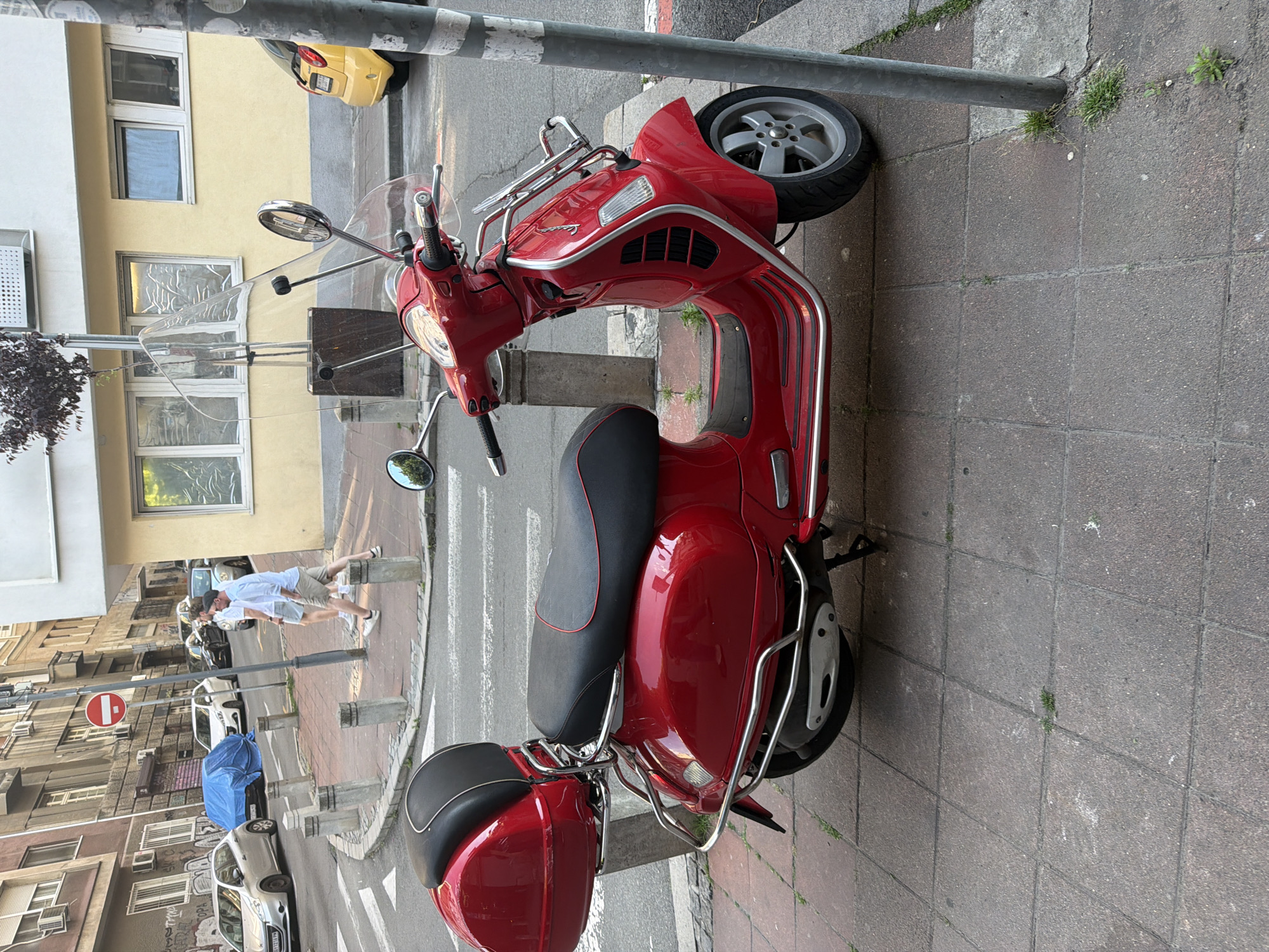 A red Vespa scooter with chrome accents is parked on a gray tile sidewalk next to a metal pole in Belgrade, Serbia. The scooter has a black seat with red trim, a clear windshield, and a luggage carrier on the back. The scooter faces right in the image. Behind the scooter is a street with cars, buildings, and pedestrians. A yellow car is parked on the street. Two people are walking on the street. A "Do Not Enter" sign is visible. The buildings are old and have windows with bars. The sky is overcast.