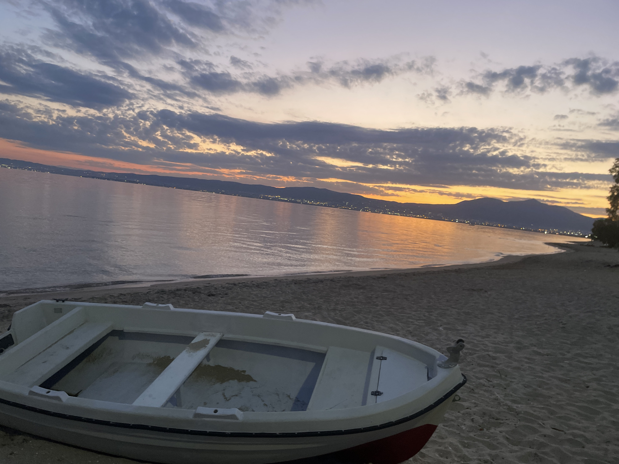 A small white boat with a red stripe rests on a sandy beach in Neoi Epivates, Greece. The boat is in the foreground, its interior showing some sand. Behind the boat, the calm sea stretches to the horizon, reflecting the warm hues of a twilight sky filled with scattered clouds. The distant coastline is illuminated by a line of lights, suggesting a town or city. The overall scene depicts a peaceful evening by the sea.