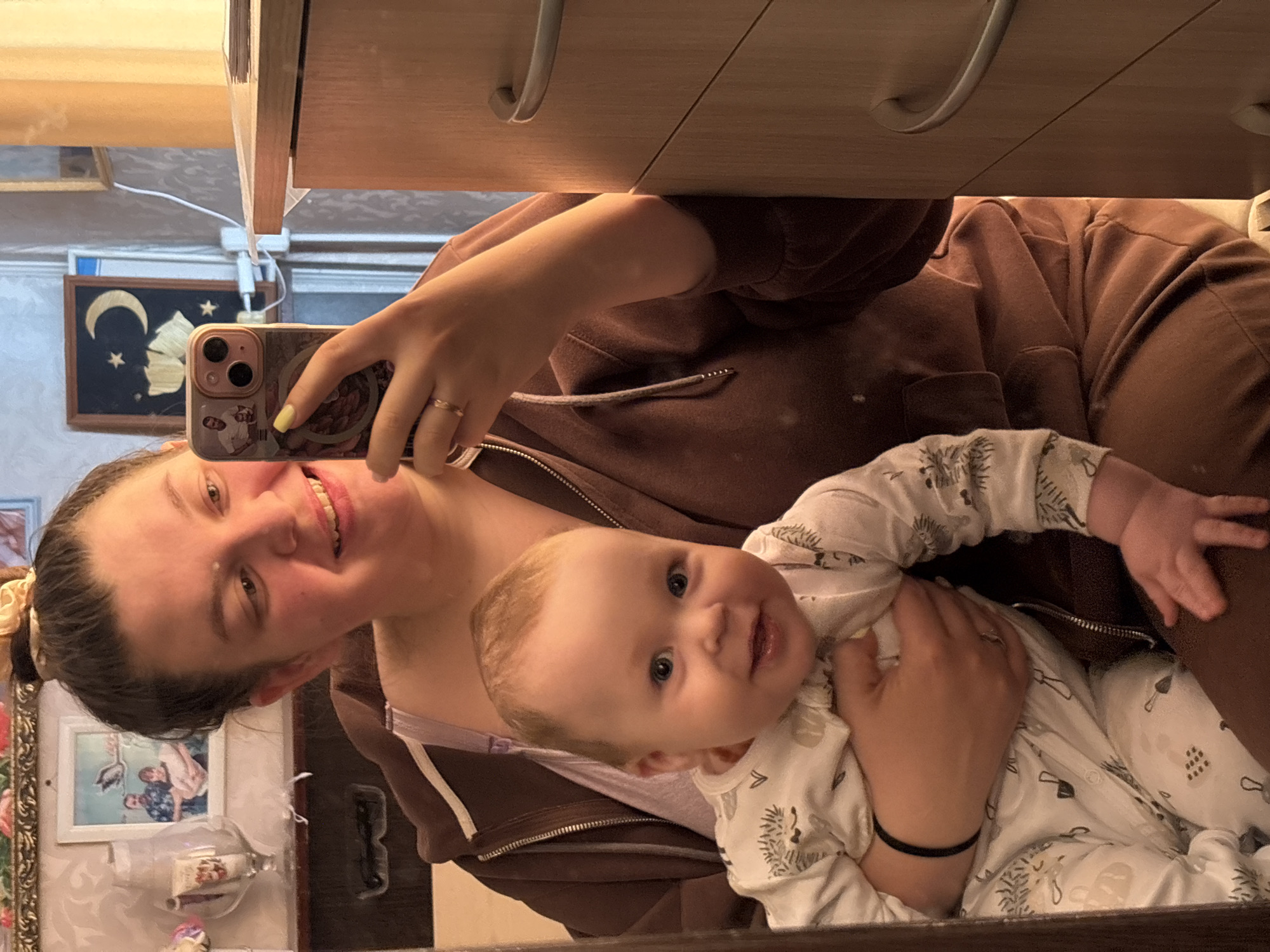 The image is a mirror selfie taken indoors, featuring a woman holding a baby. The woman, positioned on the left, is smiling broadly at the camera, her dark hair tied back with a light yellow scrunchie. She wears a brown zippered hoodie over a light-colored top. Her right hand, with light yellow nail polish and a gold-colored ring, holds a smartphone (an iPhone with a triple camera setup and a decorative case) to capture the reflection. A black elastic band is visible on her left wrist.
The baby, held by the woman in the center of the frame, has fair skin, light hair, and blue eyes, and looks calmly towards the camera with a slight smile. The baby is dressed in a white long-sleeved onesie adorned with subtle patterns, possibly depicting plants or simple tools.
The background, visible in the mirror's reflection, includes a light brown wooden cabinet or shelf above. On the wall behind the woman, there is a framed dark-colored artwork featuring a yellow crescent moon and stars. Below this, another framed picture shows two individuals. Other small decorative items and some cables are also visible, suggesting a domestic setting.
The scene captures a happy, intimate moment between the mother and child. No discernible text is visible within the image. The location context is Lukintsy, Russia.