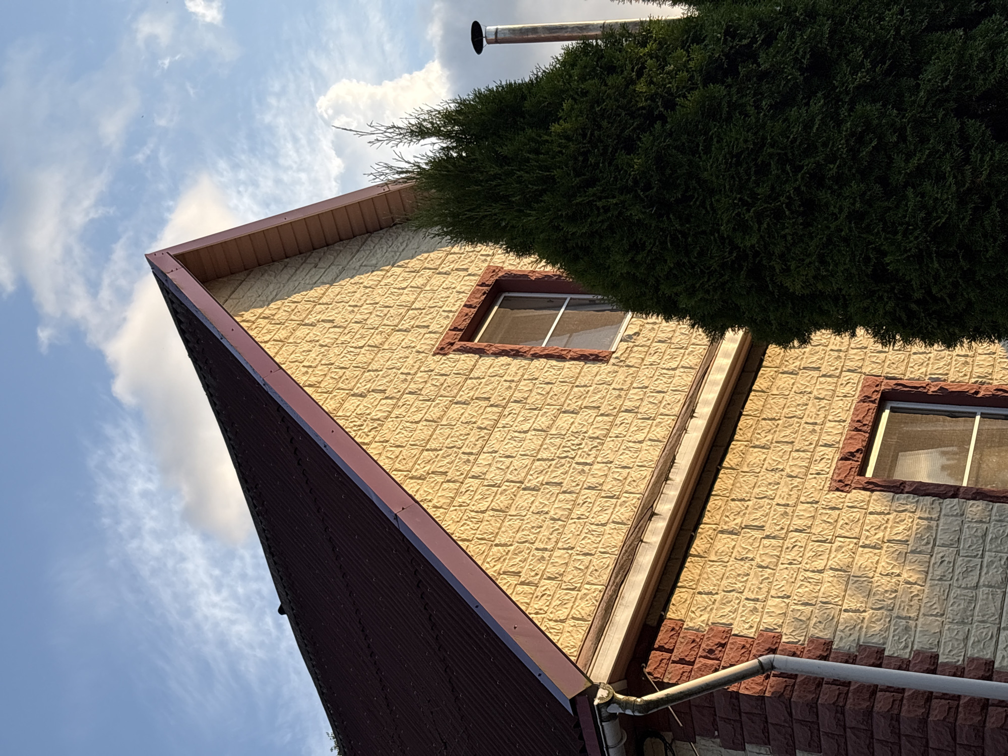 The image shows the exterior of a two-story house with a pitched, dark red metal roof. The walls are clad in two types of textured material: a light tan brick pattern on the upper portion and a darker reddish-brown brick pattern on the lower portion. There are two visible windows with red trim. A tall, dark green evergreen tree stands in front of the house, partially obscuring the right side of the structure. A silver metal chimney pipe extends upwards from behind the tree. The scene is set against a blue sky with scattered white clouds, illuminated by what appears to be late afternoon or early evening sunlight. A white downspout is attached to the corner of the house on the left. The image appears to be taken in Levashovo, Russia, based on the provided context.