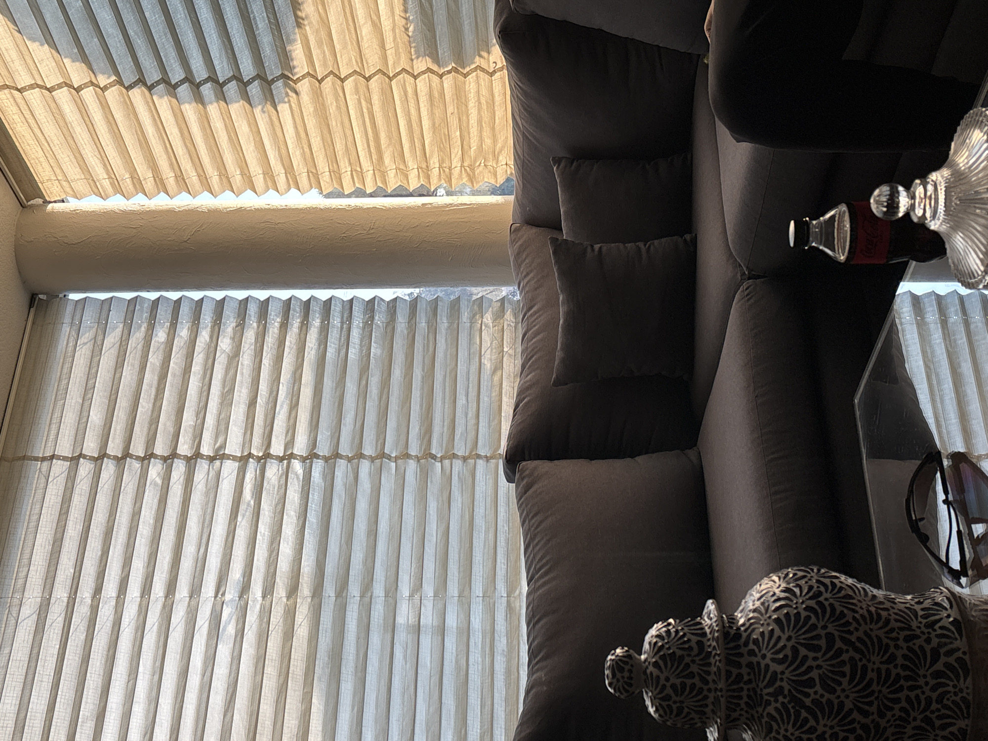 The image displays an indoor living space, captured from a slightly elevated perspective and rotated vertically. On the right side, a dark gray sofa with matching pillows is visible, partially obscured by the camera's angle. A person's arm or shoulder is faintly seen resting on the top portion of the sofa.
On a nearby surface, likely a glass table, a small glass bottle with a red label bearing the text "Coca-Cola" is placed alongside a clear, decorative glass object, possibly a vase or candle holder. Further down, a pair of sunglasses rests on the table, reflecting ambient light.
The left side of the frame is dominated by two large sections of light-colored, pleated window blinds. The upper set of blinds is brightly illuminated by strong sunlight filtering through, creating a warm glow and distinct shadows from its folds. A textured, horizontal architectural beam separates the upper blinds from a second, lower set of identical pleated blinds, which receive less direct light.
The scene suggests a daytime setting, with abundant natural light entering through the windows, indicating clear weather outside. The overall impression is one of a comfortable and well-lit interior space.