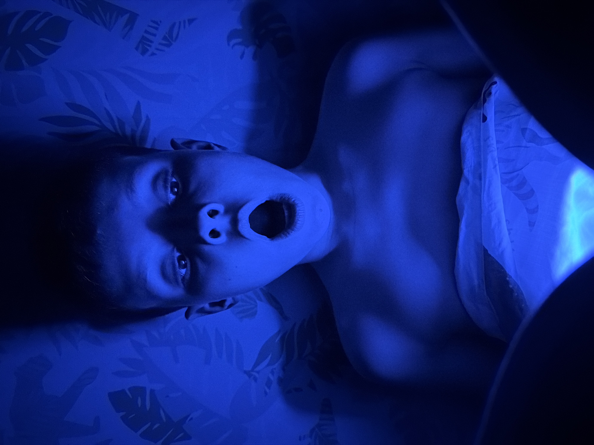 A person is lying on a patterned surface, possibly bedding with a tropical leaf print, illuminated by a strong blue light. The person's mouth is open in an expression of surprise or shock. A portion of a patterned white fabric covers their torso. The scene has a stark, dramatic lighting with deep shadows. There is no indication of a location in Votkinsk, Russia.