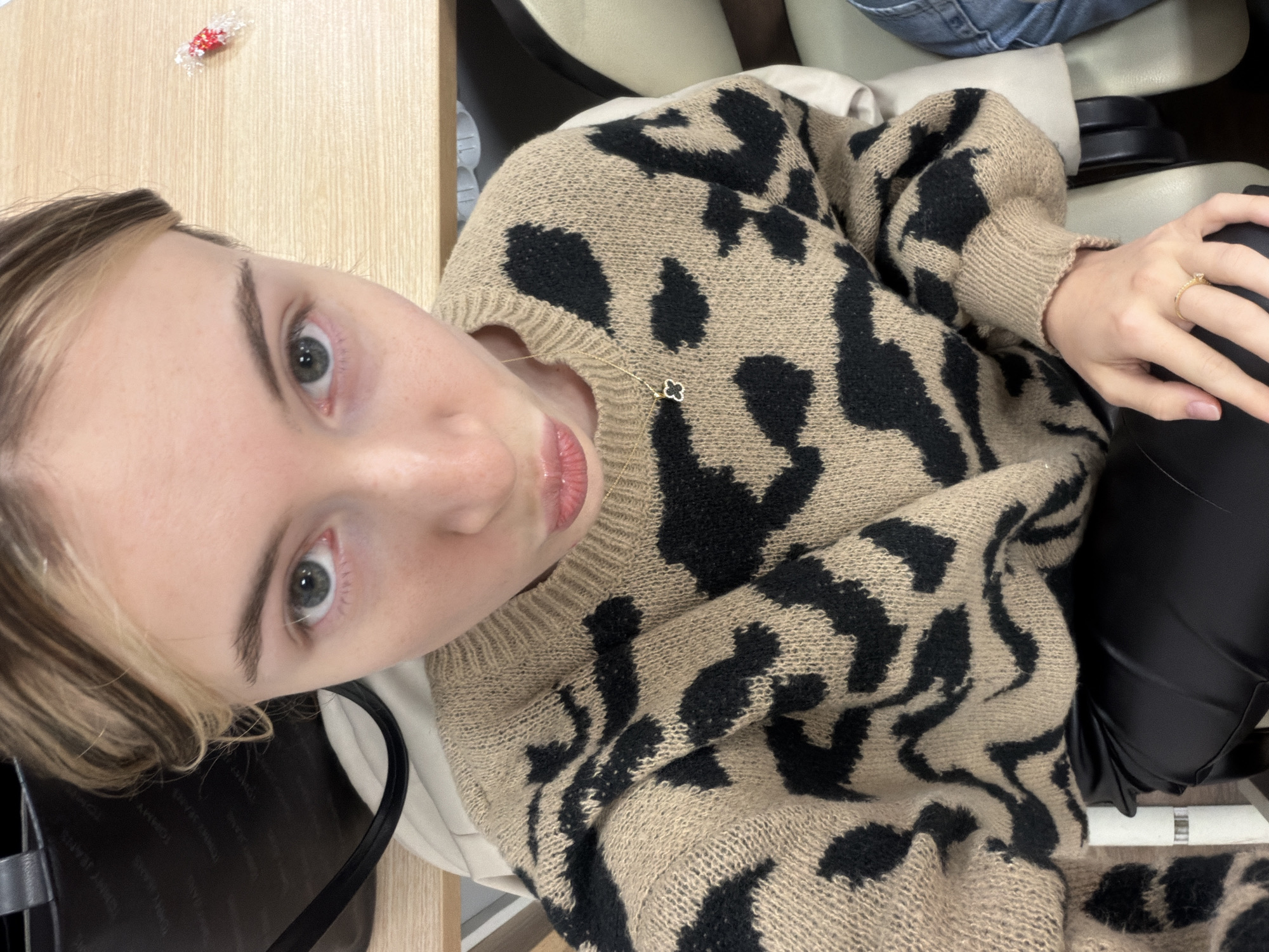 A close-up, high-angle, indoor shot shows a fair-skinned young woman with blonde hair and gray eyes, wearing a brown and black animal-print sweater. She appears to be seated, with a glimpse of a black purse or bag visible on the left side of the frame. A gold necklace with a clover pendant is visible near her neck. A gold ring is on one of her fingers. The setting appears to be indoors, possibly in a chair at a cafe or office. A wrapped piece of candy sits on the table. The exact location in Plovdiv, Bulgaria, cannot be determined from this image alone.