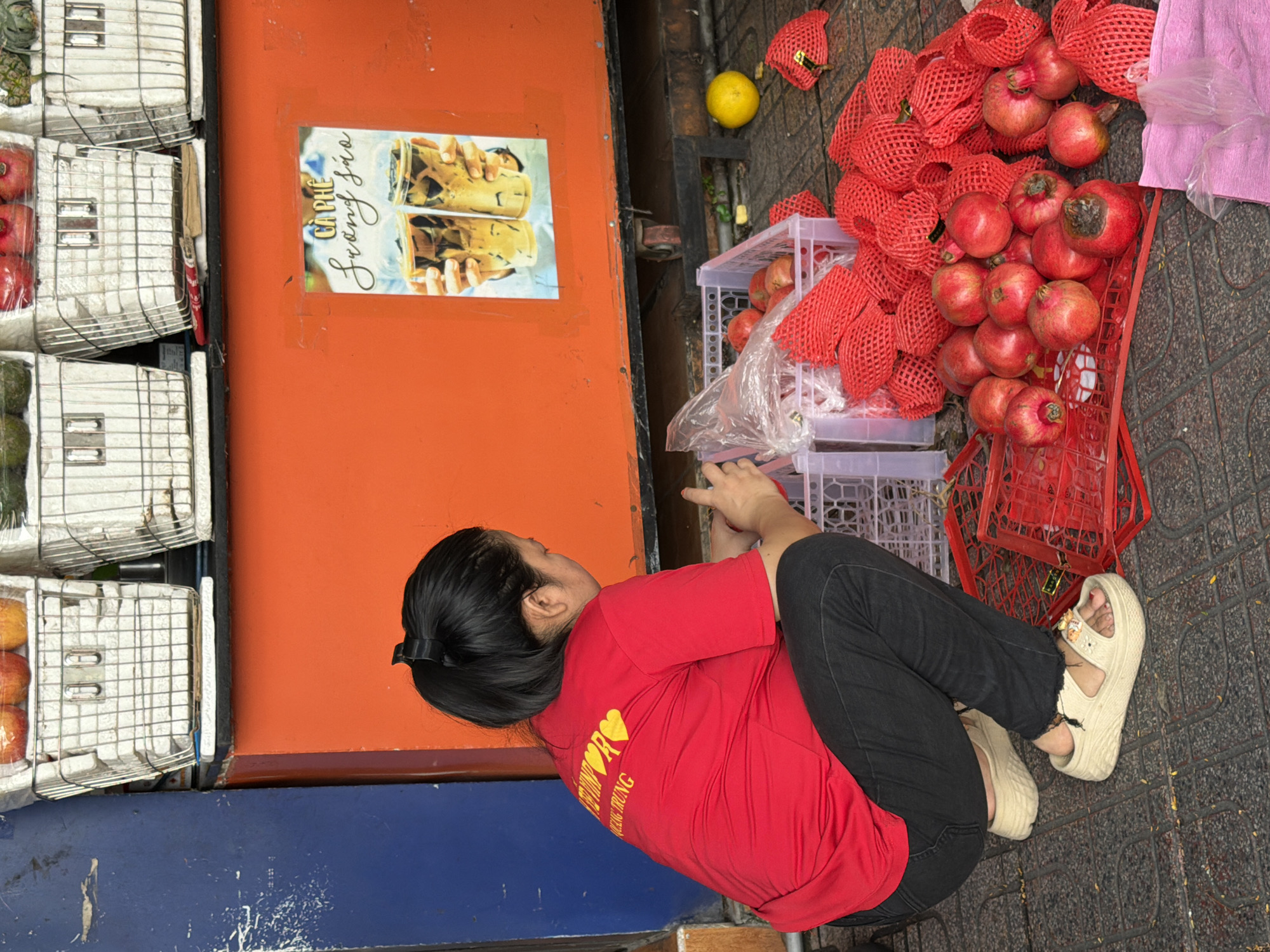 A woman in a red shirt and black pants crouches on a tiled sidewalk in Nha Trang, Vietnam, near a fruit stand. She is working with pomegranates, some of which are in red mesh netting, and others are piled on a red grate. A yellow fruit, possibly a grapefruit or pomelo, rests on the ground to the right. In the background, an orange structure displays a poster advertising coffee. Baskets of fruit are visible above the orange structure. The scene appears to be a street-side market.