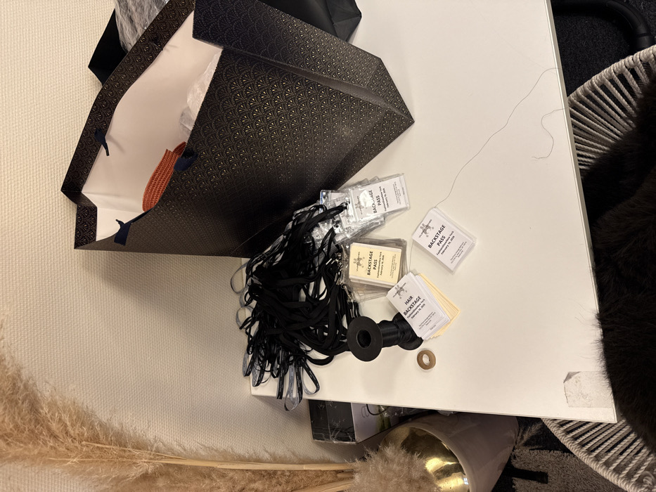 On a white surface sits a dark patterned shopping bag, partially revealing a light-colored lining and an orange garment inside. Several backstage passes labeled "February 16, 2020," along with their black lanyards, are scattered beside the bag. One spool of black ribbon is also visible with a small bronze ring. The bottom left corner shows pampas grass against a white wall. A wicker chair and a fluffy dark-colored item are on the right side.