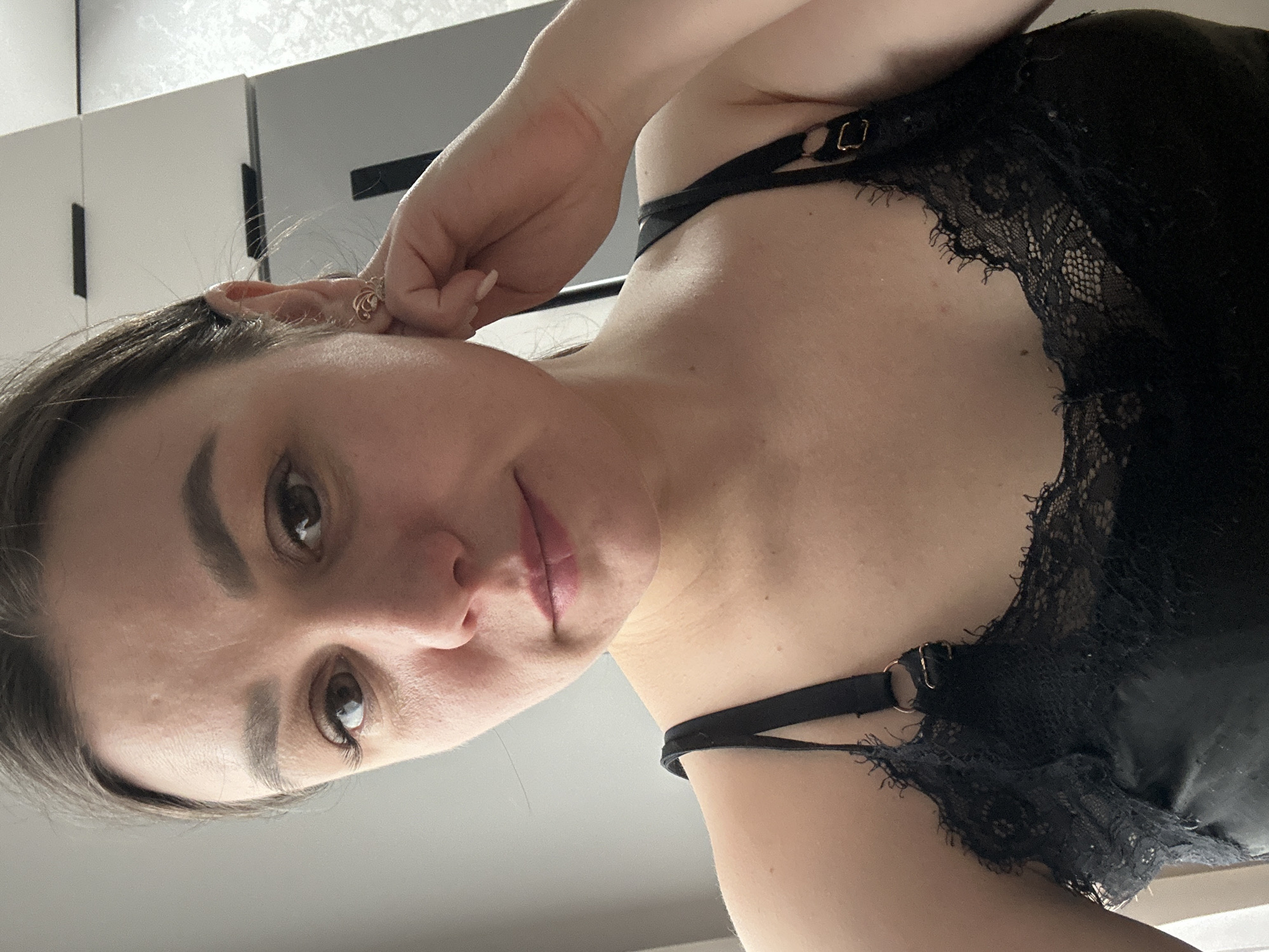 A woman is seen in a close-up, tilted perspective. She wears a black lace top with straps. Her left hand is raised near her ear, which has a ring. The background includes light-colored cabinets or walls. There is no verifiable information regarding the location being in the city of Chaikovsky, Russia from the image.