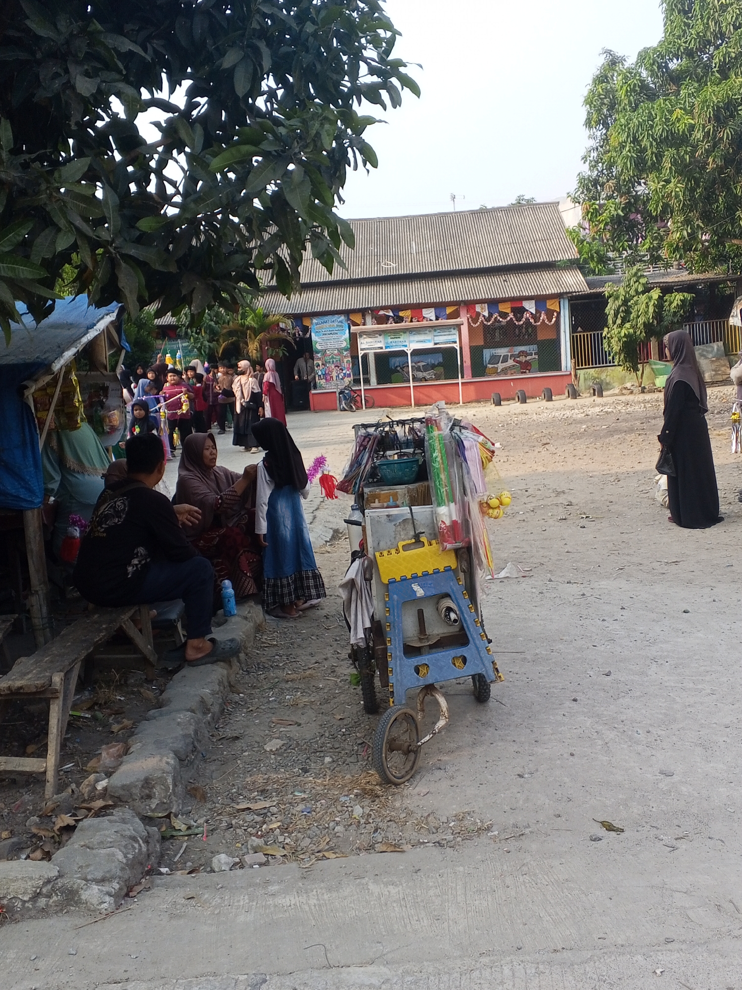 In Ciruas, Indonesia, a street scene unfolds with a group of people gathered near a building with a corrugated metal roof. A woman in a black hijab stands to the right of the frame, facing away from the camera. In the foreground, a person wearing a black t-shirt and dark pants sits on a wooden bench, facing a woman in a brown hijab and patterned skirt who is seated on the ground. A young girl in a blue skirt stands between them. Beside them is a cart filled with various items, including colorful decorations, bottles, and what appear to be toys. Behind the seated individuals, more people, some in hijabs, are visible, suggesting a communal gathering or market activity. The ground is dusty and unpaved, with scattered debris. Trees with green foliage frame the scene, and the sky is overcast. The building in the background has murals and banners displayed, hinting at a festive or commercial establishment.