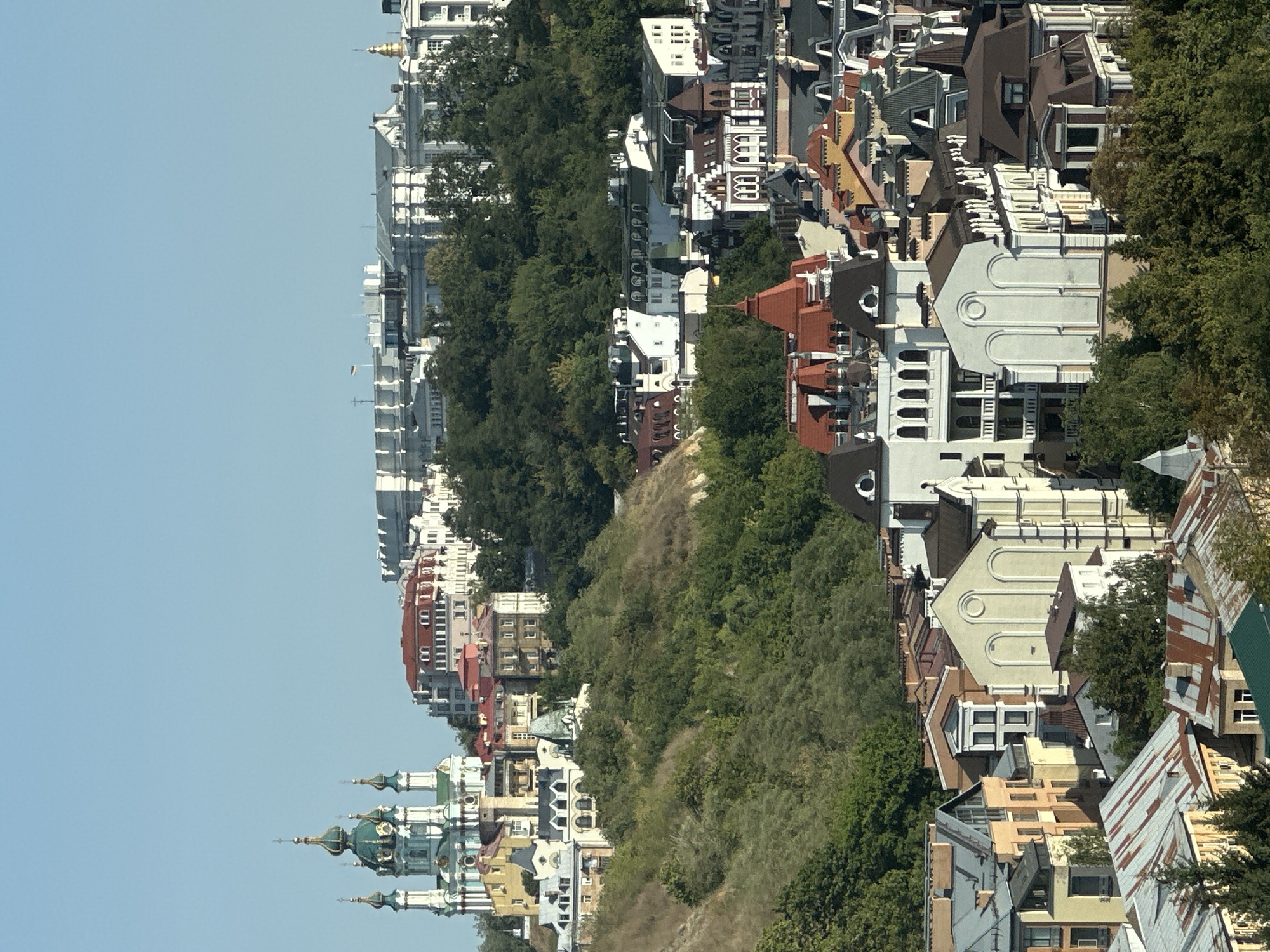 This image shows a panoramic view of Podil district in Kyiv, Ukraine. In the background, the iconic St. Andrew's Church, with its distinctive green domes, stands prominently on a hill. The church is a Baroque masterpiece, with white walls and ornate decorations.
Below the church, a cluster of colorful buildings with red and yellow roofs create a vibrant tableau. These buildings, varying in architectural styles, contribute to the charming aesthetic of the district. Further down, more buildings with diverse roof designs, including gabled and mansard roofs, extend into the foreground. Many of these structures feature white facades with intricate detailing, while others are adorned with terracotta and stone elements. The scene is nestled within a lush green landscape, with trees covering the hillsides and interspersed among the buildings. The sky above is a clear, bright blue, indicating a sunny day. The overall impression is one of a picturesque urban landscape with historical and architectural significance.