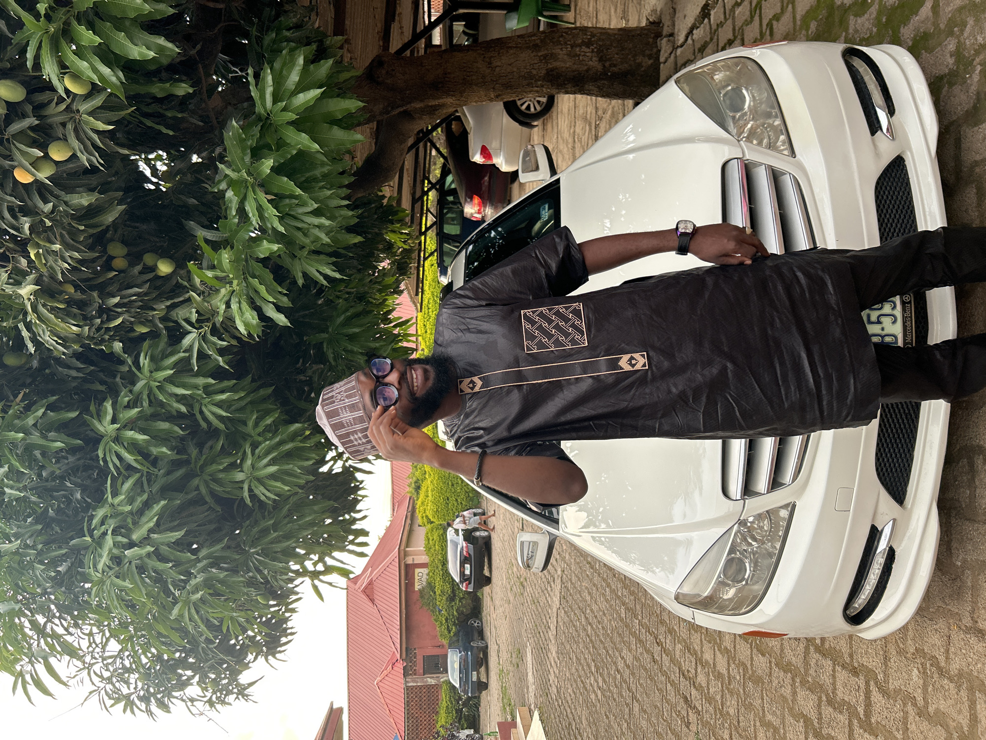 A man stands beside a white Mercedes-Benz car on a paved surface, likely a driveway, in Jos, Nigeria. He is wearing a dark-colored African-style shirt with decorative patterns, matching pants, a patterned cap, and sunglasses. He has a beard and a watch on his left wrist. His right hand is raised, near his face. A leafy tree with mangoes hangs over the scene. The background includes a building with a red roof, other parked cars, and additional greenery. The license plate of the car in front of the Mercedes-Benz is partially visible with B-59.
