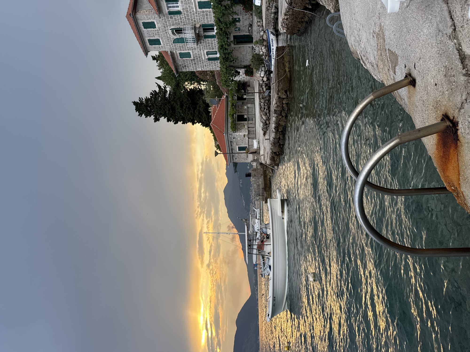 This is a landscape photograph of a coastal scene at dusk. The image is oriented vertically, with the sky at the left and the water and a stone dock at the right. In the foreground on the right, a metal ladder extends into the water from the dock. The dock is made of rough, grey stones and concrete.
Behind the dock, a body of water is visible, reflecting the warm colors of the sunset. Several boats are moored near the dock, including a white motorboat with visible text that reads "ZL 154".
Further back, on the left side of the image, traditional stone buildings with red-tiled roofs are nestled against a backdrop of dark green trees and mountains. The sky on the left is a pale grey, transitioning to hues of orange and yellow where the sun is setting behind the mountains. The overall mood is serene and tranquil, suggesting a peaceful evening in a Mediterranean-like setting. The lighting indicates it is either sunrise or sunset.