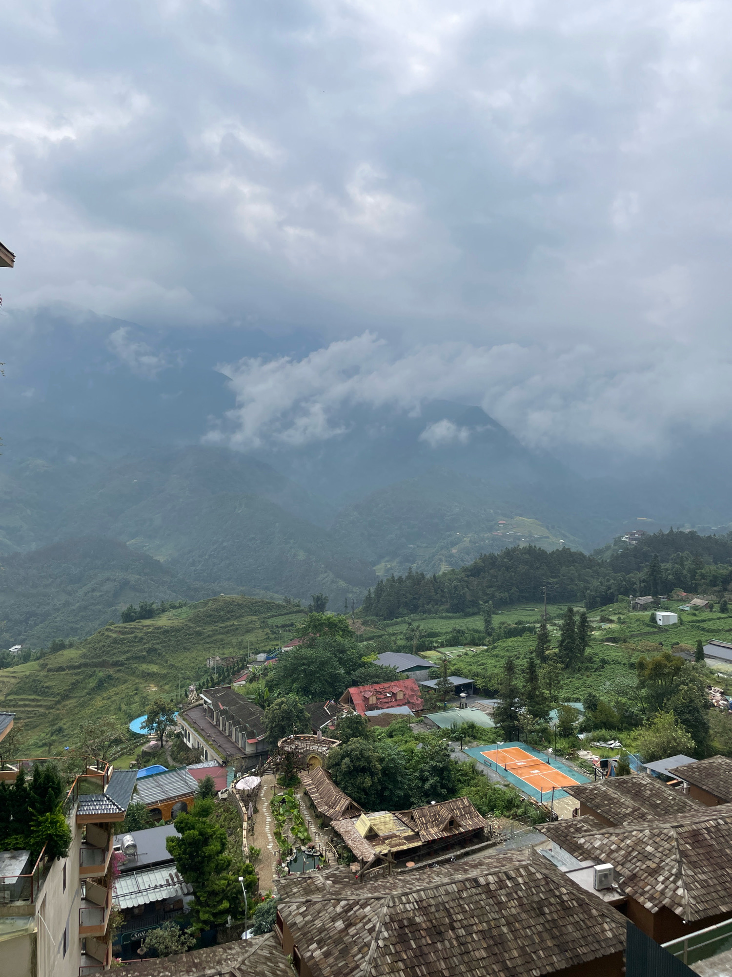 The image depicts an aerial view of a village nestled in the mountainous region of Sa Pa Ward, Vietnam. The landscape is characterized by lush green terraced hillsides and dense foliage, interspersed with various buildings. In the foreground, several structures with distinct tiled roofs are visible, suggesting a residential or resort area. Further into the mid-ground, a bright orange tennis court with blue trim stands out against the surrounding greenery, indicating recreational facilities. A collection of buildings with a mix of architectural styles, including some with darker tiled roofs and others with red roofs, are spread across the terrain. The background is dominated by rolling hills and mountains, partially shrouded in a soft, overcast sky with scattered clouds. The overall impression is a serene, rural setting with a blend of natural beauty and human habitation.
