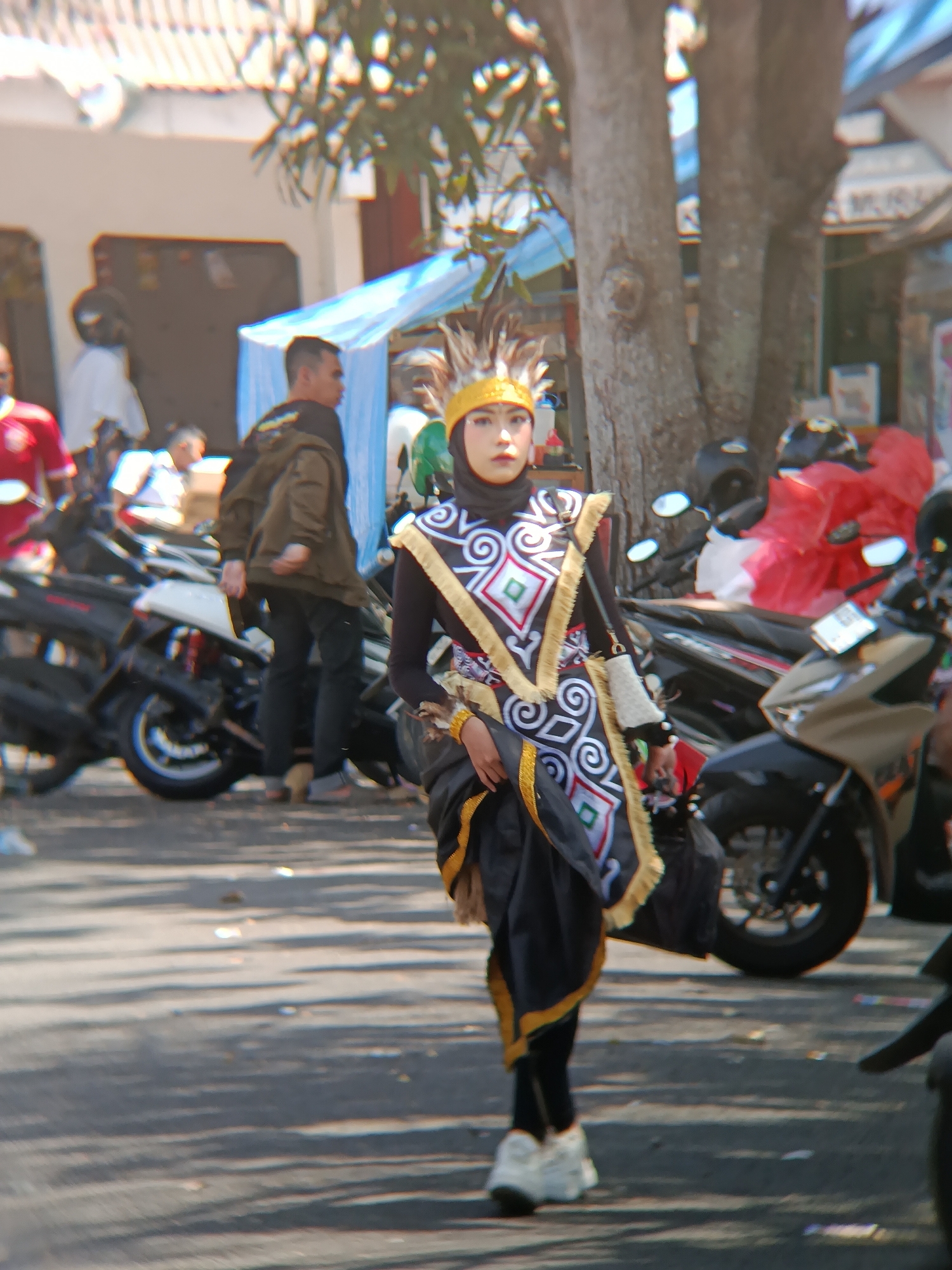 A young woman in traditional Indonesian attire is walking on a paved street in Desa Kebon Manis, Kecamatan Cilacap Utara, Indonesia. Her costume consists of a black hijab, a black long-sleeved shirt, and a black skirt with gold trim. Over the shirt, she wears a decorated vest with white, green, and red diamond patterns, along with gold fringe. A feathered headdress adorns her head. She is wearing white sneakers. The scene appears to be outdoors during the daytime, with trees and parked motorcycles in the background. Several other individuals are present, some walking and others near the motorcycles. A white tent-like structure is visible in the background.
