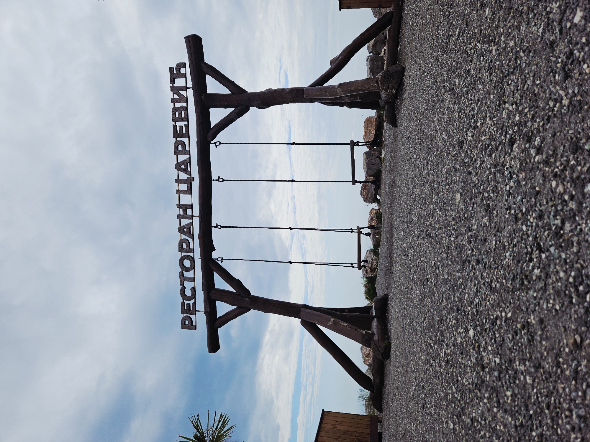 The image shows a large wooden sign in the city of Unknown, Unknown. The sign reads "РЕСТОРАН ЦАРЕВИЋ" in Cyrillic script, suggesting a restaurant named "Tsarevich". The sign is constructed from rough-hewn logs and features three swing seats suspended from the main structure. The ground is covered in gravel. The sky is visible in the background with a mix of blue and white clouds. A palm tree and a wooden structure can be seen in the lower left and lower right corners respectively.