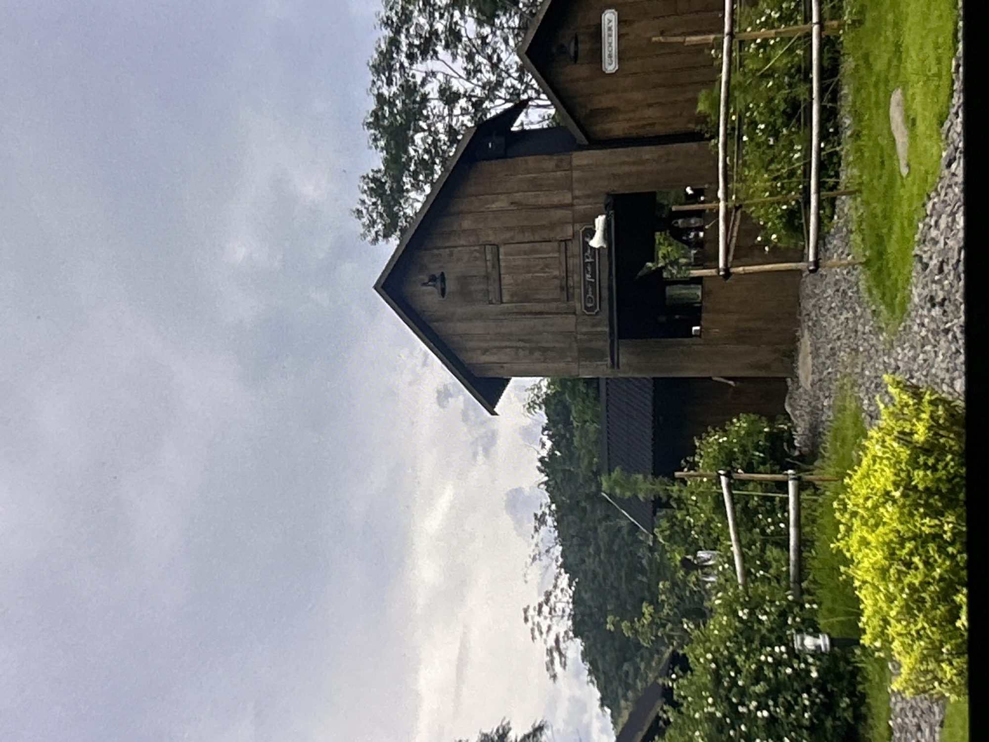 The image depicts a rustic wooden building complex, potentially a farm or a themed attraction, set against a backdrop of lush greenery and a cloudy sky. The main structure is a two-story wooden barn-like building with a steeply pitched roof. A smaller, attached structure to the right has a sign that reads "GROCERY." A white object, possibly a ceramic figurine or a small animal, rests on a ledge above an open entryway of the main building. The foreground features a gravel pathway leading towards the buildings, bordered by a low wooden fence and manicured green grass. Bushes with white flowers are visible throughout the scene. The overall atmosphere suggests an outdoor rural setting, possibly within the city of Depok, Indonesia, given the mention of its location.