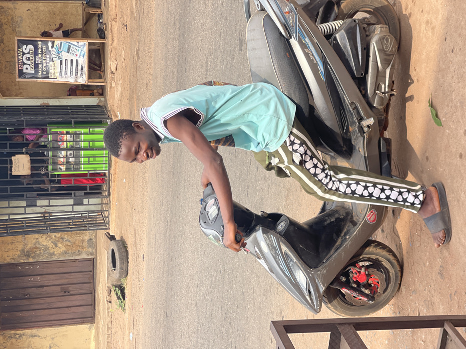 An outdoor scene in Neni, Nigeria, depicts a young adult male seated on a dark gray scooter, positioned slightly off the asphalt road on a dirt shoulder. The male, of African descent, wears a light blue sleeveless top with striped trim, olive green track pants featuring a distinct white and black geometric pattern down the sides, and dark slides. He is smiling and looking towards the camera, with both hands on the scooter's handlebars. The scooter displays a visible Kymco logo on its side and has a red front brake caliper.
In the background to the left, a light brown building with a large closed brown metal rolling door is visible. A discarded tire lies on the ground in front of it, along with some sparse vegetation. Further right, behind the male subject, a storefront is secured by a dark metal security gate. Inside, two individuals are partially visible: one in a pink top and another in red clothing, near a bright green display unit. A small, handwritten sign hangs from the gate.
To the far right, a white A-frame sign advertises "EZENWATA POS AVAILABLE" and "CHARGE YOUR PHONE & OTHER DEVICES HERE," detailing services such as money transactions and bill payment. A small child is partially visible near the base of this sign, adjacent to a wooden bench. The overall scene indicates a commercial street environment under daylight conditions.