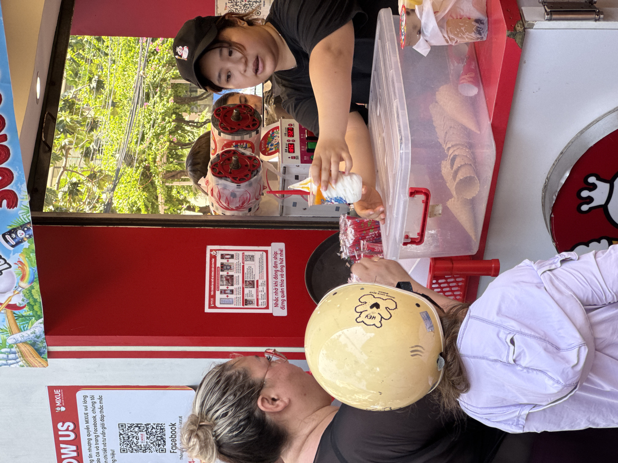 This image captures an outdoor Mixue ice cream and tea stall in Nha Trang Ward, Vietnam, during daylight. A young Asian female vendor, wearing a black cap featuring a white character and a black t-shirt, is positioned behind the red and white counter. She is actively extending a clear plastic cup filled with white soft-serve ice cream topped with orange syrup towards a customer.
The primary customer, an adult female with blonde-streaked hair styled in a bun and red-framed glasses, stands partially with her back to the camera, looking at the vendor. In front of her, a child, viewed from the back, wears a light purple hooded jacket and a yellow helmet adorned with a "Hey GOOD MILO BEAR" graphic and a bear outline.
The stall's counter features a large clear plastic container filled with numerous waffle cones. A red basket holds individually wrapped red items, likely straws or spoons. Behind the vendor, red and white machines, potentially soft-serve dispensers, display digital readings such as "10.5C" and "132.1". The stall's red panel displays Vietnamese text detailing Mixue's product packaging standards and a reminder for delivery orders to include spoons and straws. A white sign on the adjacent wall presents "FOLLOW US" information for Mixue's social media, including a QR code for Facebook. The lower part of the stall features the Mixue brand's white "Snow King" mascot logo. The background reveals lush green trees and numerous overhead electrical cables, indicating an urban outdoor setting. Another individual's face is partially visible behind the vendor.