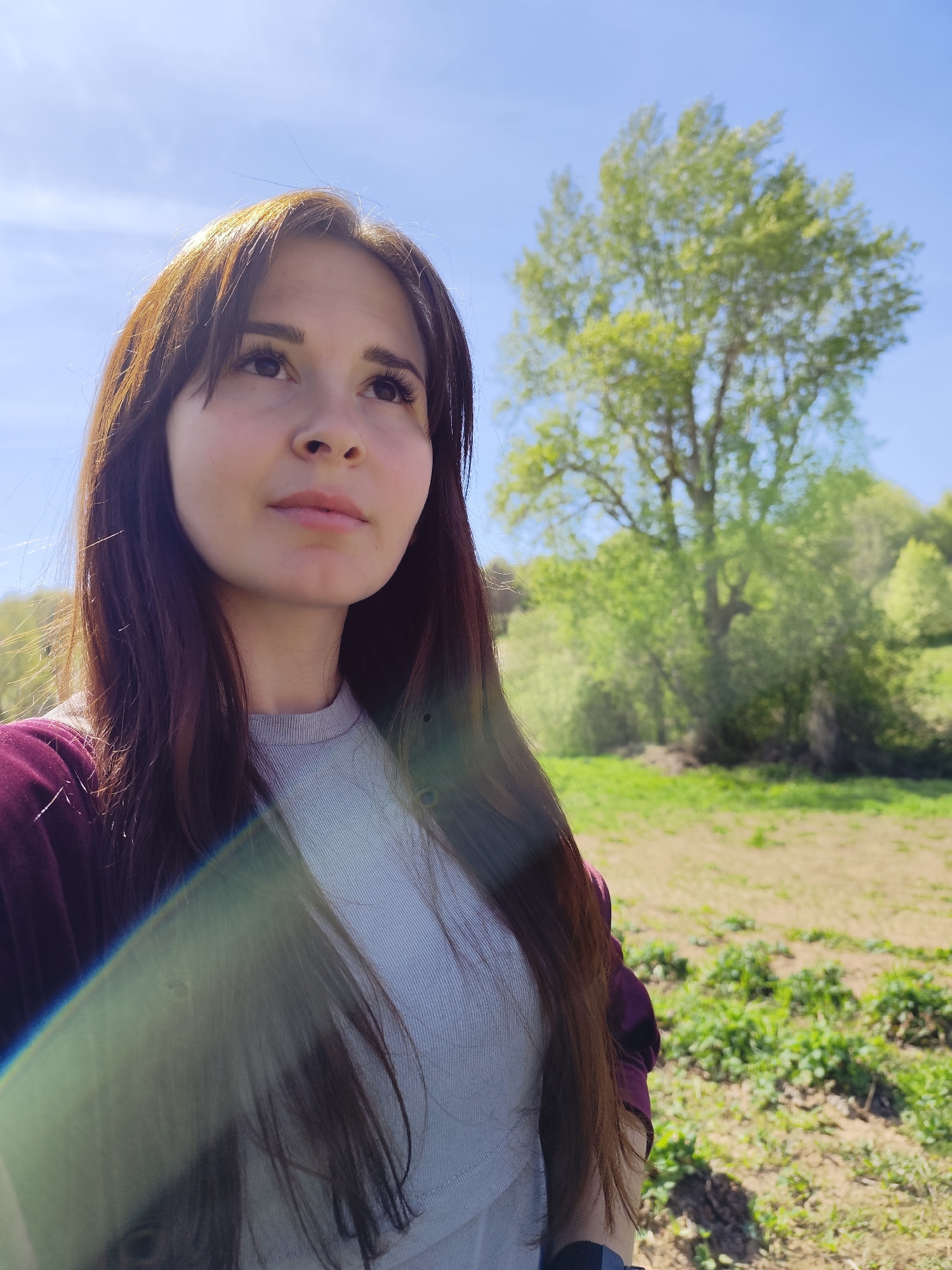 A young woman with long brown hair and bangs is standing in an outdoor setting. She is wearing a light gray ribbed shirt and a dark purple jacket. She is looking slightly upwards. The background consists of a grassy field with some patches of vegetation, and a tree with green leaves is visible in the distance. The sky is blue with a few clouds. A light flare is visible near the bottom left corner of the image. The scene appears to be taken in natural daylight. There is no information to suggest the location is in the city of Фоки, Russia.