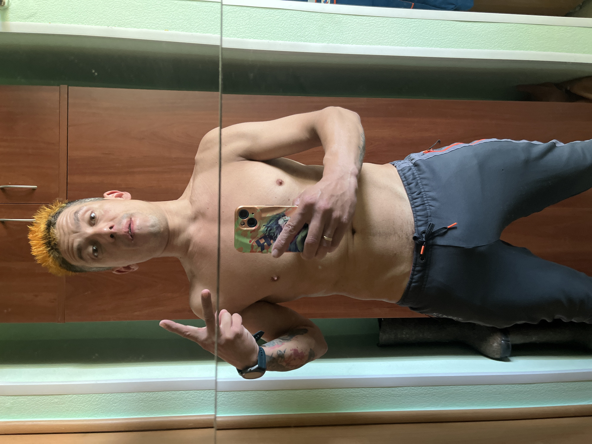 A shirtless man is seen in a mirror selfie, wearing grey sweatpants with orange accents, holding a phone with a green and brown patterned case, and giving a peace sign. He has short, orange-colored hair. He appears to be in a bathroom, indicated by the mirror, cabinet, and vanity area. The cabinets are a light brown wood. The wall is painted a light green. A dark object, possibly footwear, is on the vanity surface. There is no visible information in the image that indicates this photo was taken in Chaikovsky, Russia.