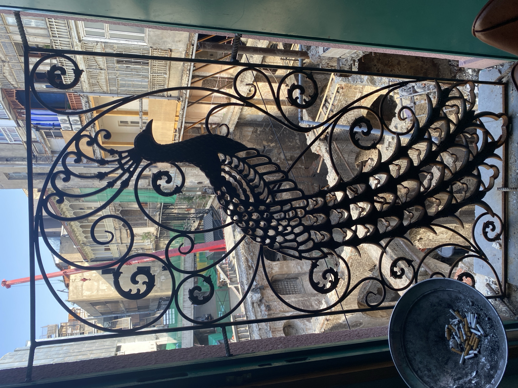 An outdoor scene is viewed through a decorative wrought-iron gate featuring a peacock silhouette, looking down into an urban courtyard. The courtyard appears to be undergoing renovation or demolition, with exposed brickwork, concrete structures, and construction materials visible. A large red crane is prominent in the background, extending upwards against a clear blue sky. Multiple buildings with balconies and windows, some appearing aged and others with air conditioning units, surround the courtyard. In the foreground, to the left, a metal ashtray is filled with cigarette butts and ash. The overall impression is one of an urban environment, likely in Tbilisi, Georgia, given the architectural style and the presence of construction.