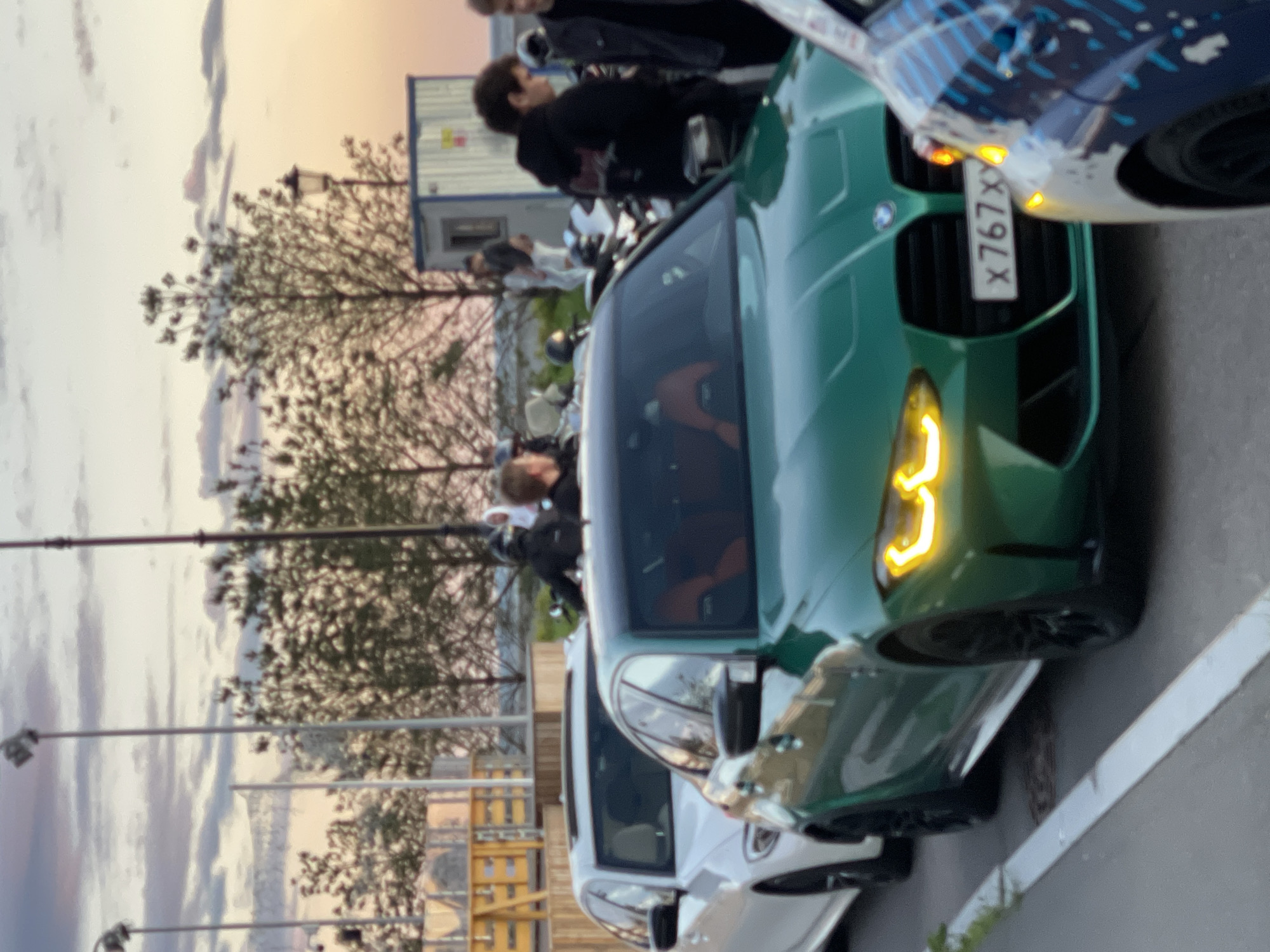 An outdoor scene at dusk features several cars and people. In the foreground, a green BMW M3 with its headlights illuminated is parked on a road, partially obscuring a white car to its left. The BMW's license plate is visible as "x767xx". To the right of the green BMW, a portion of another vehicle with a blue and white design is visible. In the middle ground, several people are standing and interacting near motorcycles. Behind them, trees with sparse foliage are visible against a dusky sky with scattered clouds. A light pole with a spotlight is positioned on the left side of the image. In the background, a temporary structure resembling a portable cabin is present. The overall environment suggests an urban or semi-urban setting during twilight.