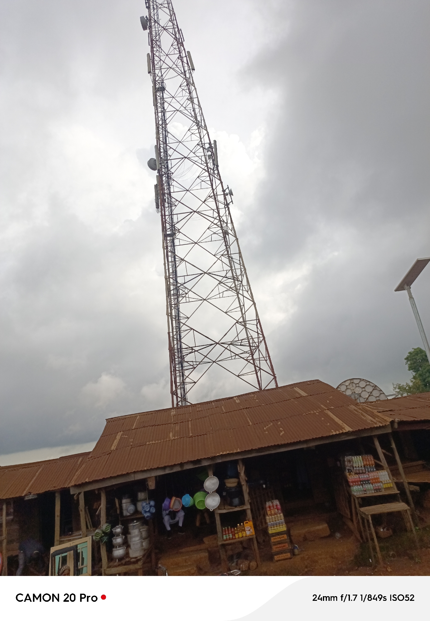 A telecommunications tower looms over a cluster of rustic buildings in Nguroje, Nigeria. The tower, constructed of metal lattice, extends high into a cloudy sky, dotted with several antennas. Beneath the tower, corrugated metal roofs, weathered to a rusty brown, cover a collection of shops. These open-fronted establishments display various goods, including buckets, cookware, and bottled beverages, suggesting a bustling marketplace. A solar-powered street lamp is visible to the right of the tower.