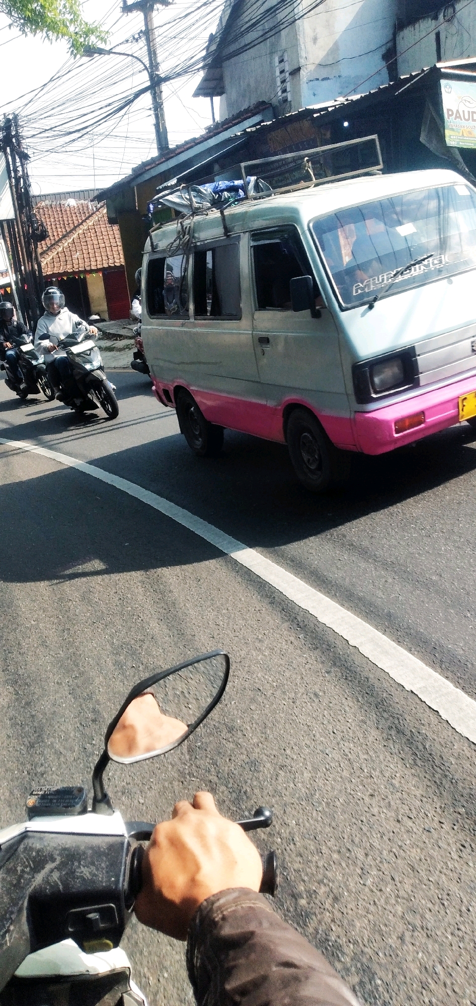 The image captures a street scene in Cilaku, Indonesia, from the perspective of a motorcyclist. The foreground shows a hand gripping the handlebar of a white scooter, with a partial view of the scooter's front. In the middle ground, two motorcycles are visible, one ridden by a person wearing a white hoodie and a helmet, and another behind it. To the right, a light blue and pink van is driving on the road, with visible lettering on its windshield. The background features buildings with tiled roofs, dense overhead wiring, and trees, suggesting a dense urban or semi-urban environment. The road has a white dividing line, and shadows are cast by the vehicles and overhead structures. The overall impression is of traffic on a sunny day.