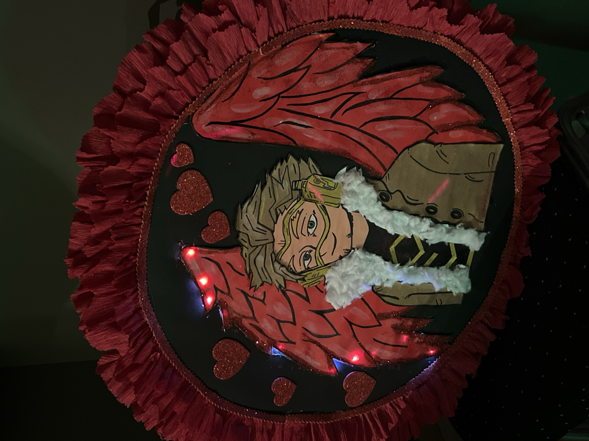 The image depicts a decorated circular object with an anime-style drawing of a character. The drawing includes red wings, a blond-haired figure with a furry collar, and various heart-shaped decorations. There are also red lights embedded in the object. The background is dark, and the location appears to be indoors. The city is unknown.