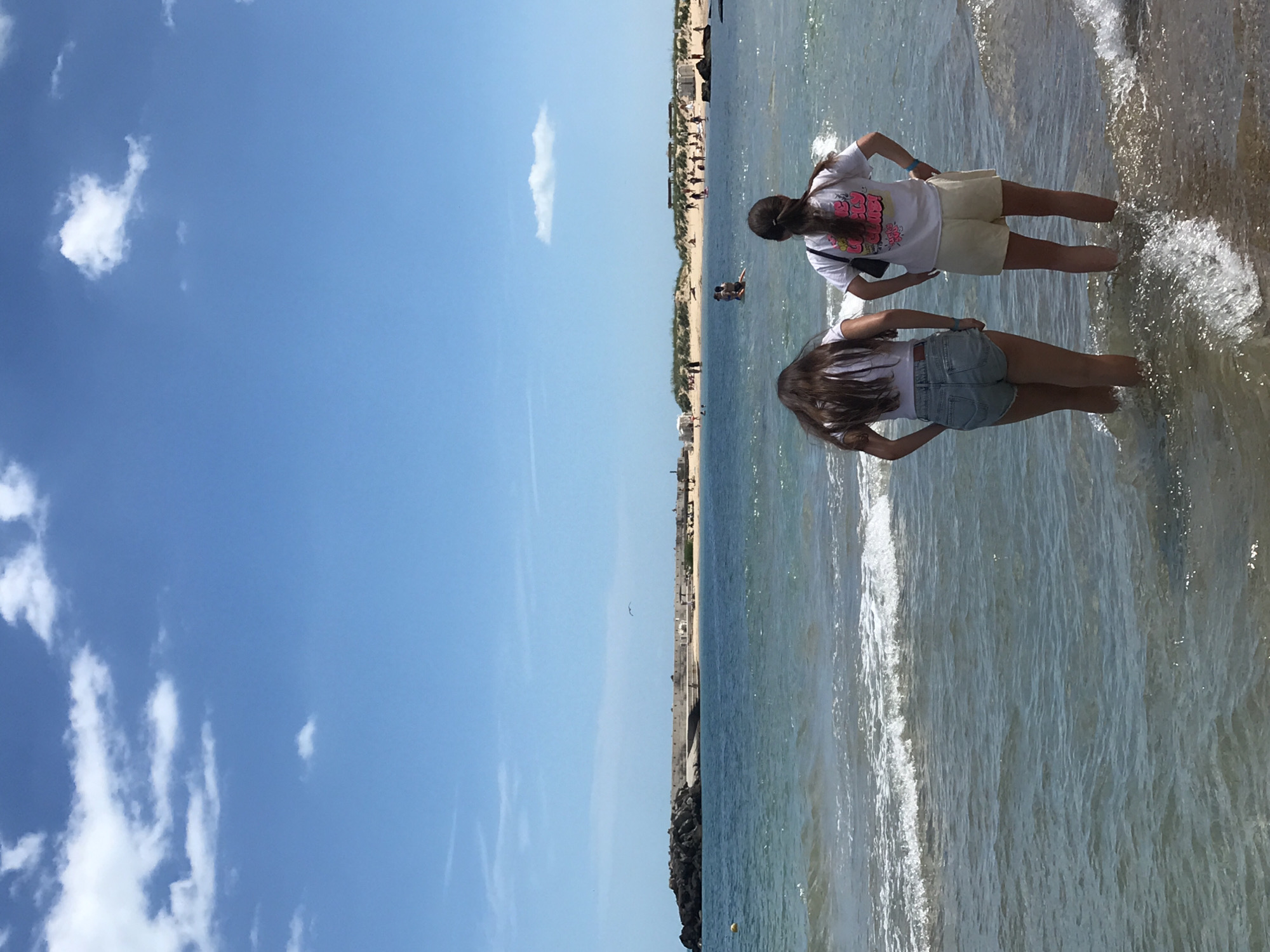This image depicts a sunny day at a beach, likely in Varna, Bulgaria, given the architectural style of the breakwater. In the foreground, two young women are standing in shallow water, their backs to the camera. The woman on the left wears denim shorts and a white t-shirt, while the woman on the right wears light-colored shorts and a white t-shirt with pink lettering. Both have their hands on their hips.
The middle ground shows the sea with gentle waves and further out, a breakwater extends from a rocky outcrop. A few people are visible on the sandy area behind the breakwater and on the beach. The background features a clear blue sky with scattered white clouds and a distant bird in flight. The overall scene conveys a relaxed, summery atmosphere.