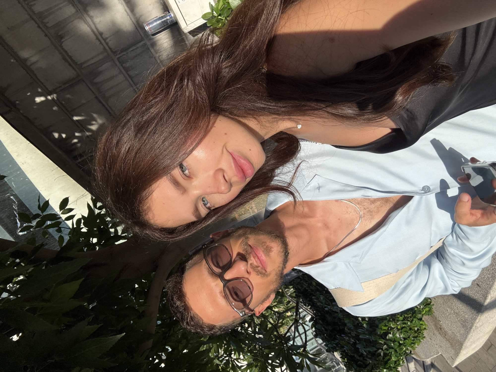 This is a selfie of a young couple outdoors in Tel Aviv, Israel, on a sunny day. The woman, with long, dark brown hair, is on the right side of the frame, her face close to the camera, with her head tilted slightly. She is wearing a black tank top and a delicate necklace with a single diamond pendant. The man, on the left, is wearing sunglasses and a light blue open-collar shirt with a beige strap visible across his chest. He has short, dark hair and a beard. He is holding a smartphone in his hand. Behind them, greenery from trees and bushes is visible, as well as a textured wall and what appears to be the edge of a building with a can of soda on a shelf. The sunlight casts distinct shadows on the wall and the people, indicating it's likely late afternoon.