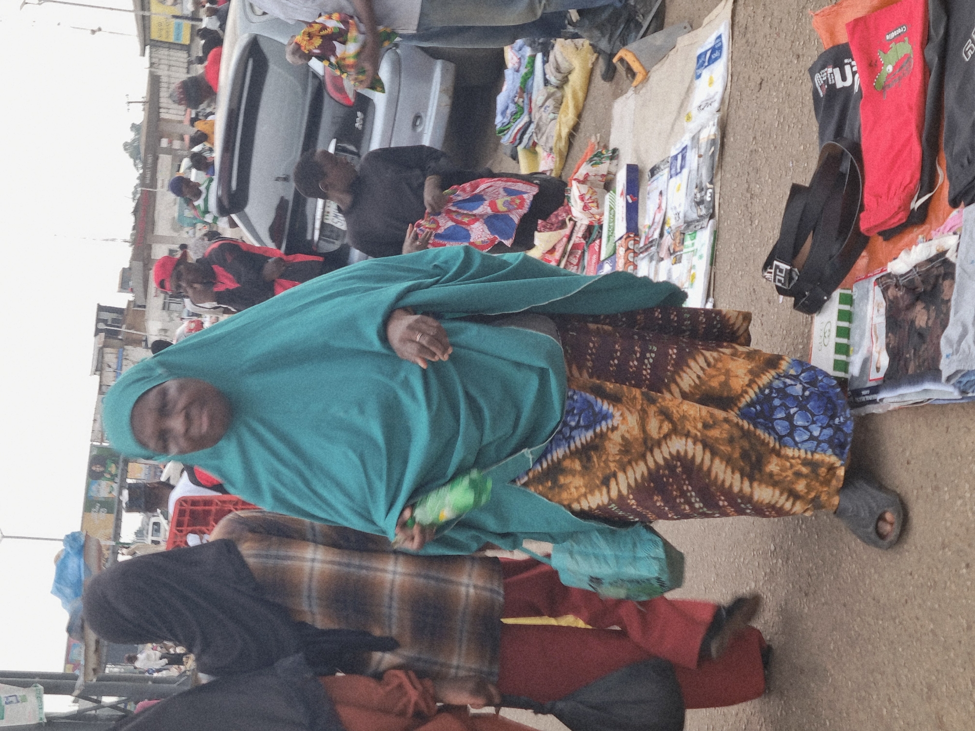 This image shows a street scene in Jos, Nigeria, likely in the morning or afternoon given the daylight. The setting appears to be a busy marketplace or roadside stall, with people and various goods displayed.
In the foreground, a person wearing a teal headscarf and a patterned skirt is prominently featured. Their hand, adorned with a ring, is visible. Beside them, another person is dressed in black pants and a red top. Further in the background, a man in a black shirt and red cap is visible near a silver car. There are also other individuals, some partially obscured, and a red crate.
The ground is paved with asphalt, and various items are spread out for sale or display. These include clothing, such as t-shirts with graphics like a crocodile on a red shirt, as well as folded fabric or blankets with intricate patterns. Some of these items are laid out on the ground, while others appear to be in bags or bundles. A green bottle is also visible.
The overall impression is one of a bustling, everyday scene in a Nigerian urban environment. The people appear to be engaged in their daily activities, possibly as vendors and customers in a market setting. There is no indication of any specific event or emotion beyond the typical activity of commerce. The weather appears to be clear, allowing for good visibility.