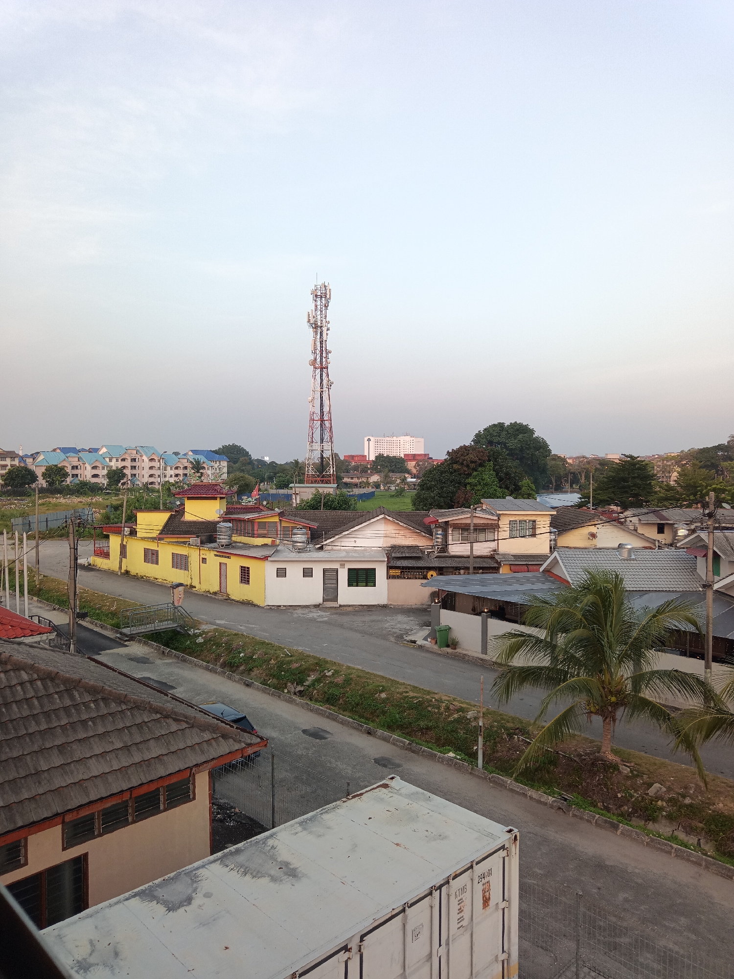 The image captures an outdoor scene in Klang City, Malaysia, characterized by a mix of residential buildings, infrastructure, and natural elements under a clear sky.
Dominating the background is a tall, red and white telecommunications tower, a prominent piece of urban infrastructure. In the middle ground, a cluster of single-story residential buildings are visible. Several of these houses are painted yellow with red tiled roofs, while others are white or beige. Some of these buildings feature water tanks on their roofs. Beyond these, a row of apartment buildings with blue roofs are situated, suggesting a more densely populated area further in the distance.
A paved road runs through the scene, with a black car parked on the side. Green vegetation, including grass and trees, lines the roadside and surrounds the buildings, with a noticeable palm tree in the foreground on the right. A white shipping container is partially visible in the lower right corner of the image, suggesting a connection to logistics or storage. The overall environment appears to be a suburban or semi-urban residential area. The lighting suggests it might be early morning or late afternoon due to the soft, diffused light.