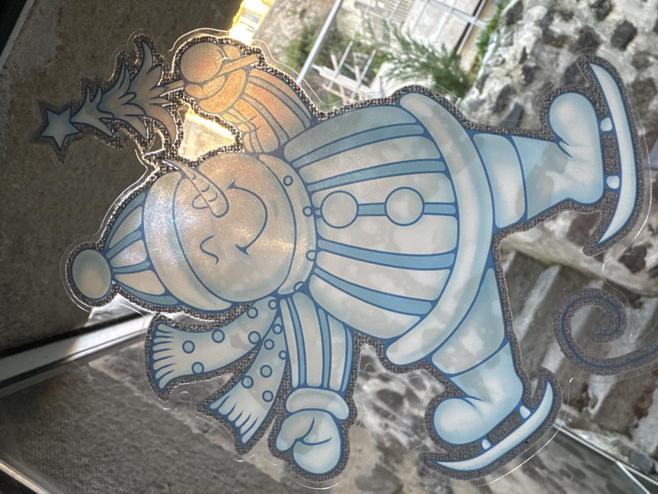 A transparent window cling, depicting a light blue and white snowman with a glitter outline, is affixed to a clear glass pane. The snowman, presented in a dynamic pose, wears a striped winter hat and a polka-dotted scarf. Its face features a carrot nose, two dots for eyes, and a wide, upturned smile. Its torso is vertically striped with three circular buttons. The snowman's raised right arm holds a stylized Christmas tree topped with a star, while its left arm is bent. It wears ice skates, with a spiral trail visible beneath the left skate, indicating movement. Through the window, an out-of-focus exterior scene reveals a textured grey surface to the upper left and center, and green foliage with a metal railing to the right. The scene is illuminated by natural light and is located in the city of Le Puy-en-Velay, France.