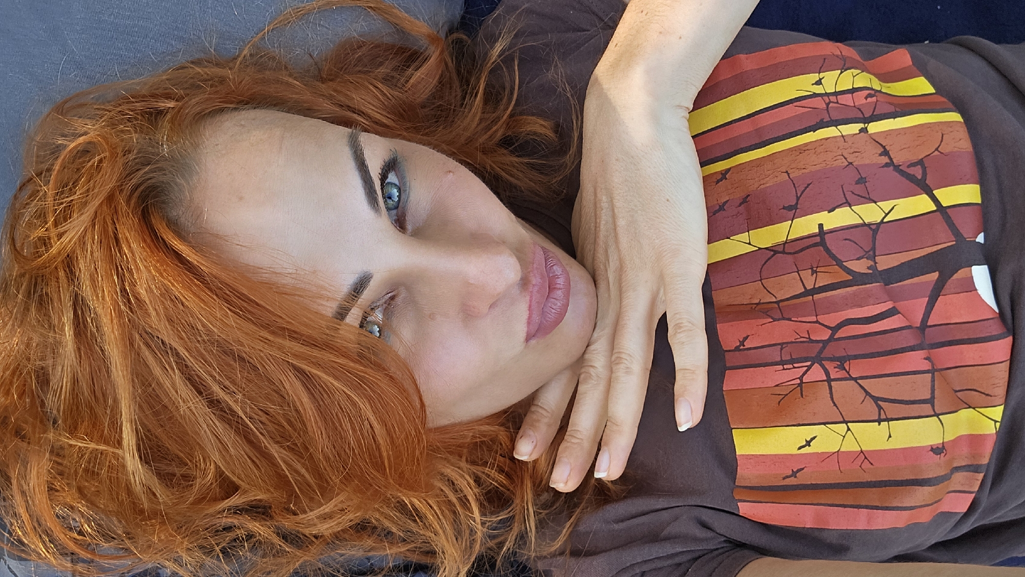 A woman with vibrant red hair and blue eyes is captured in a close-up, overhead shot. She is wearing a dark brown t-shirt with a graphic print featuring vertical stripes in shades of yellow, orange, and red, overlaid with a silhouette of bare trees and birds. Her left hand is resting on her chin, with her fingers spread. Her lips are slightly parted, and she is looking directly at the camera. The background appears to be a dark, textured surface, possibly upholstery. The image does not contain any contextual clues that would allow for the determination of its location in Warsaw, Poland, or any city.