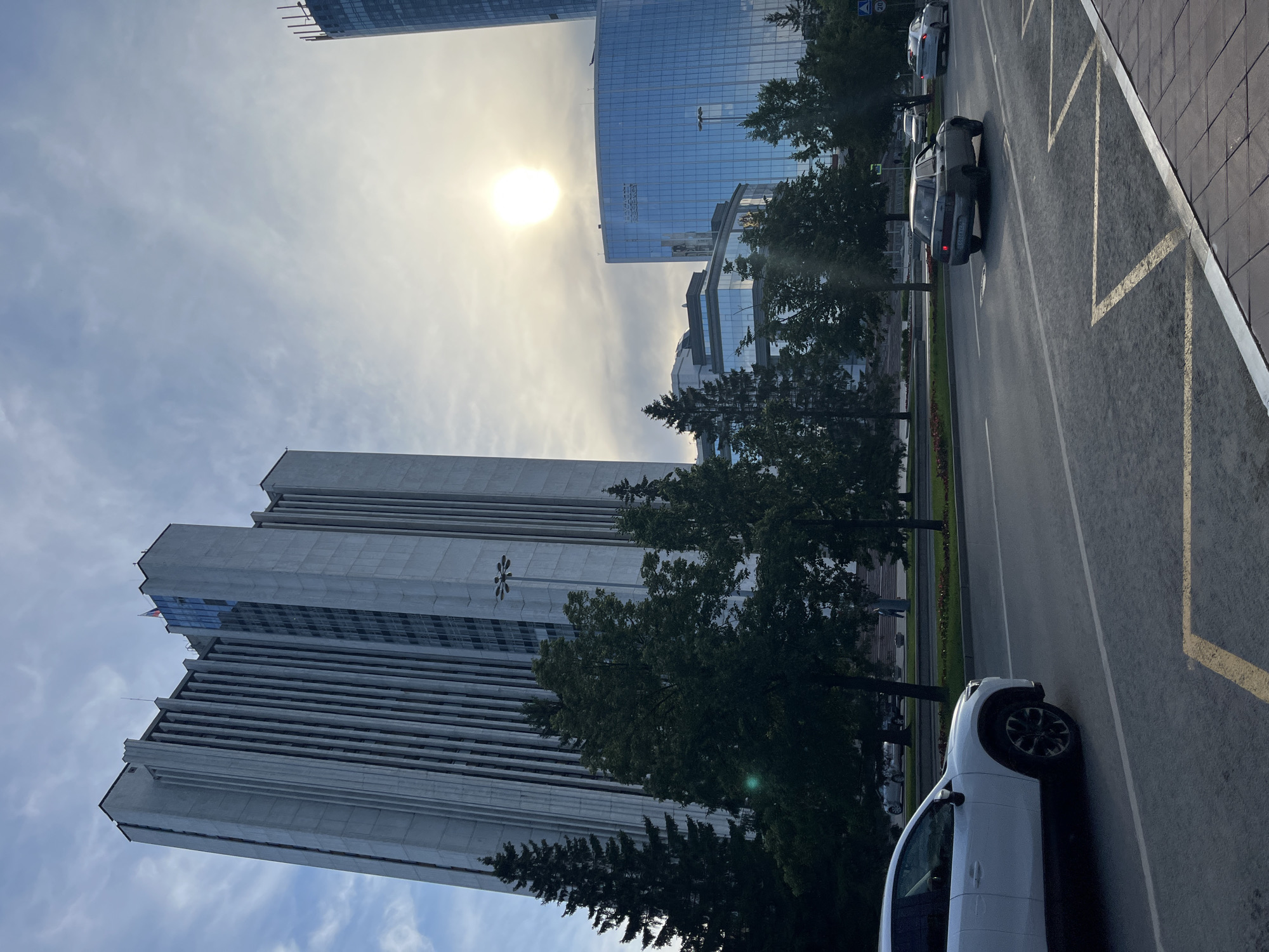 In this scene captured in the city of Unknown, Unknown, a vast expanse of sky dominates the upper portion of the frame, with a bright sun partially obscured by clouds, casting a soft glow. The sky transitions to a lighter blue near the horizon. Prominently featured are several large, modern buildings. To the left, a tall, grey skyscraper with vertical ribbed detailing and a flag on its upper section stands out. To the right, a blue glass-clad skyscraper rises, its facade reflecting the sky and surrounding environment. In front of these imposing structures, a line of lush green trees provides a natural contrast. Below the trees, a wide, paved road runs horizontally across the image. Several cars are visible on the road, including a white SUV in the foreground on the left, and two sedans in the middle ground, one dark-colored and another lighter colored further down the road. The road is marked with white lines and some yellow painted markings. The environment appears to be an urban setting with a mix of architecture and greenery.