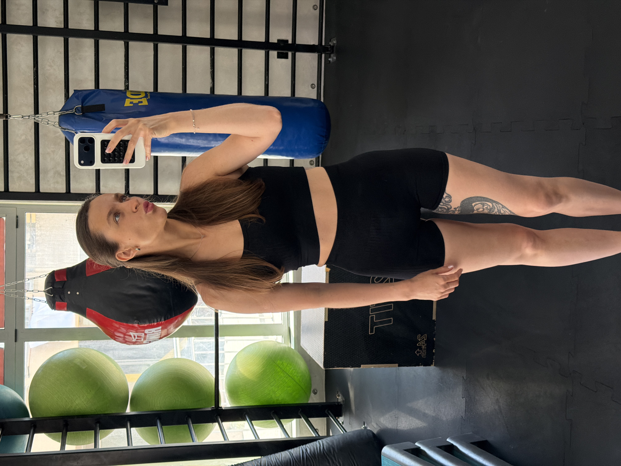 A young adult female is captured in a mirror selfie within a gym located in Budva, Montenegro. She stands centrally, facing her reflection while holding a white iPhone with a black textured case in her right hand, positioned near her face. Her lips are pursed in a pout, and her gaze is directed to her right. Her long, dark brown hair is styled straight, with some strands pulled back from her face. She wears a black ribbed athletic crop top and matching high-waisted athletic shorts. A thin silver chain with a small cross pendant is visible around her neck, and a delicate silver bracelet adorns her right wrist. A prominent black and grey tattoo of an anatomical heart and scrollwork is visible on her left thigh.
The gym environment features dark grey interlocking rubber floor mats. To the left, a black metal shelving unit holds three bright green exercise balls, with more partially visible above and below. A tear-drop shaped punching bag, predominantly black with red accents and white branding, hangs from the ceiling near a large window. Through the window, blurred architectural details of an urban setting are visible. To the right, a blue cylindrical punching bag with yellow Cyrillic lettering (partially visible as "БУД...") hangs, positioned in front of black wall bars mounted on a light grey wall. A black plyometric box with white lettering is situated on the floor behind the female.
