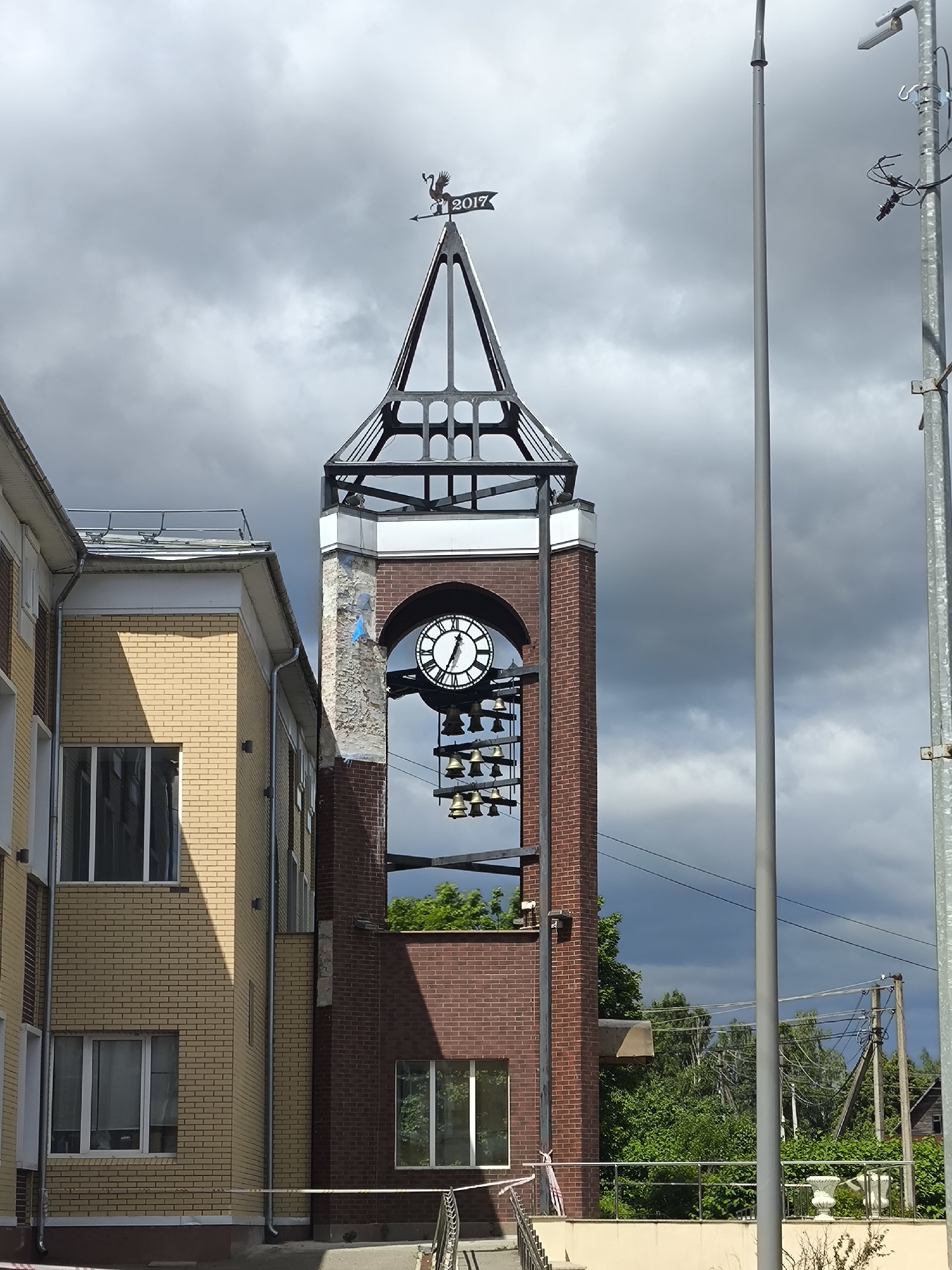 A clock tower with bells and a weather vane is shown in Taldom, Russia. The tower has a brick facade and is topped by a metal structure with the year "2017" visible on the weather vane. The clock face is round with black numerals and hands. Below the clock face, multiple rows of bells are suspended. Adjacent to the clock tower is a yellow brick building with windows. In the background, there are trees and power lines. The sky is overcast.