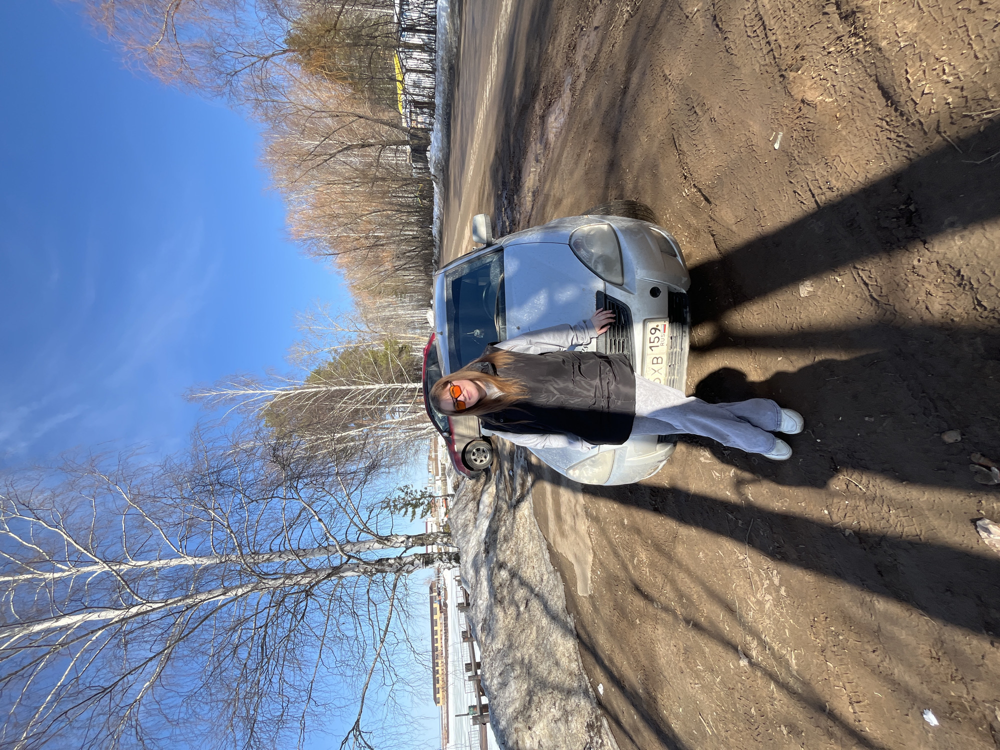 The image captures an outdoor scene in Chaikovsky, Russia, under clear blue skies, indicative of early spring or late winter conditions. A female individual stands centrally, leaning with her right arm against the front-left fender of a silver Lada sedan. She is wearing a black sleeveless puffer vest over a light grey long-sleeved top, light grey pants, and white sneakers, with orange-tinted sunglasses on her face. The sedan displays a Russian license plate "9X B 159 RUS" on its front bumper.
The ground directly in front and to the left of the car is a muddy, brown dirt surface with distinct tire tracks and long, dark shadows extending from the left, suggesting a sunny environment. To the left of the dirt area are mounds of dirty, melting snow and ice, bordering a partially frozen body of water. Across this water, in the background, multiple low-rise structures with yellow and brown facades are visible. A partial view of a red sedan is present behind and to the left of the silver car. To the right, an asphalt road runs parallel to the dirt shoulder. The environment is characterized by numerous bare deciduous trees, predominantly birches, and some evergreen trees lining the background along the road and water's edge.